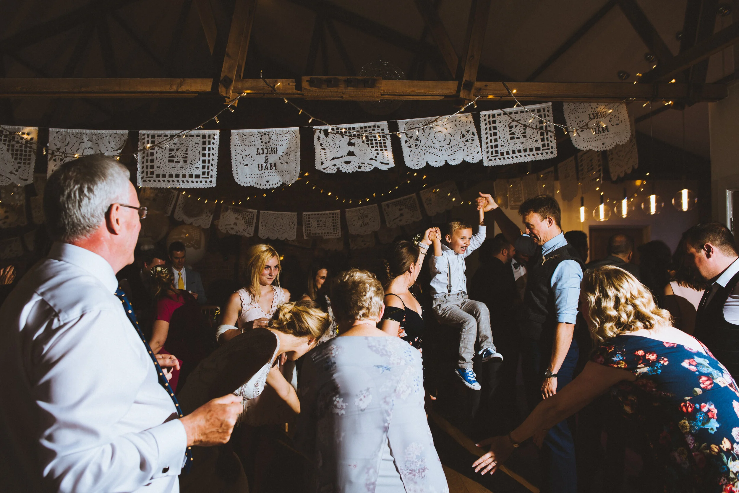 People dancing and celebrating at a festive gathering with papel picado banners and string lights overhead.
