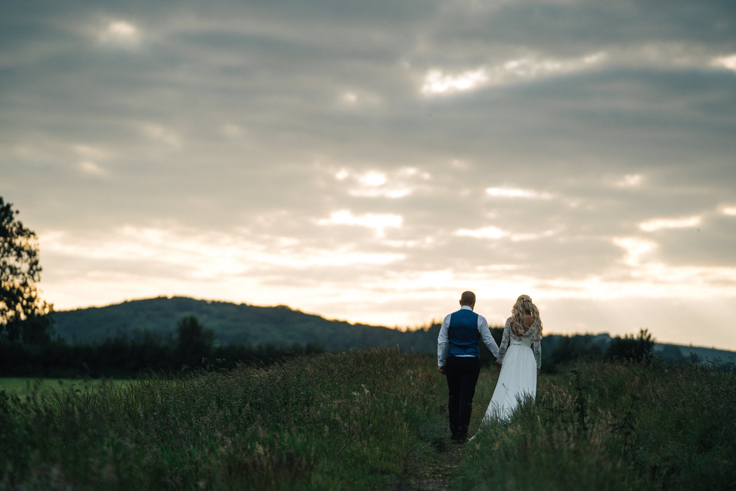 A couple in wedding attire walking hand in hand on a grassy path through a field, with hills in the distance and a cloudy sky overhead.