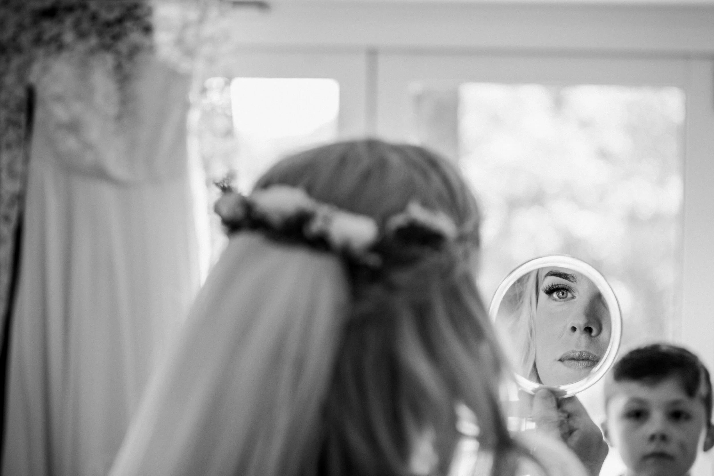 A woman holding a mirror reflects her face, showing eye makeup and lips, while a young boy stands nearby.