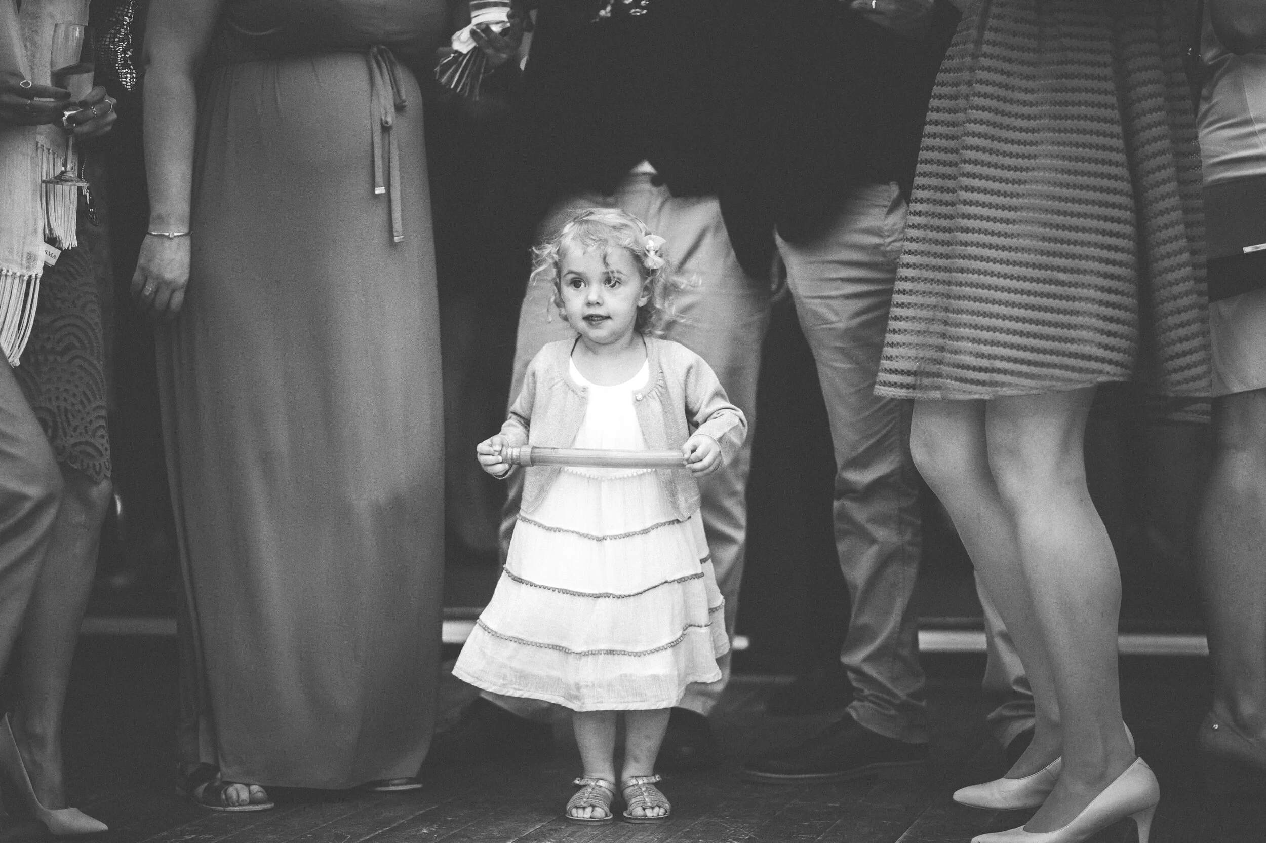 A young girl with curly hair holding a stick, standing between adults in a crowded indoor setting.