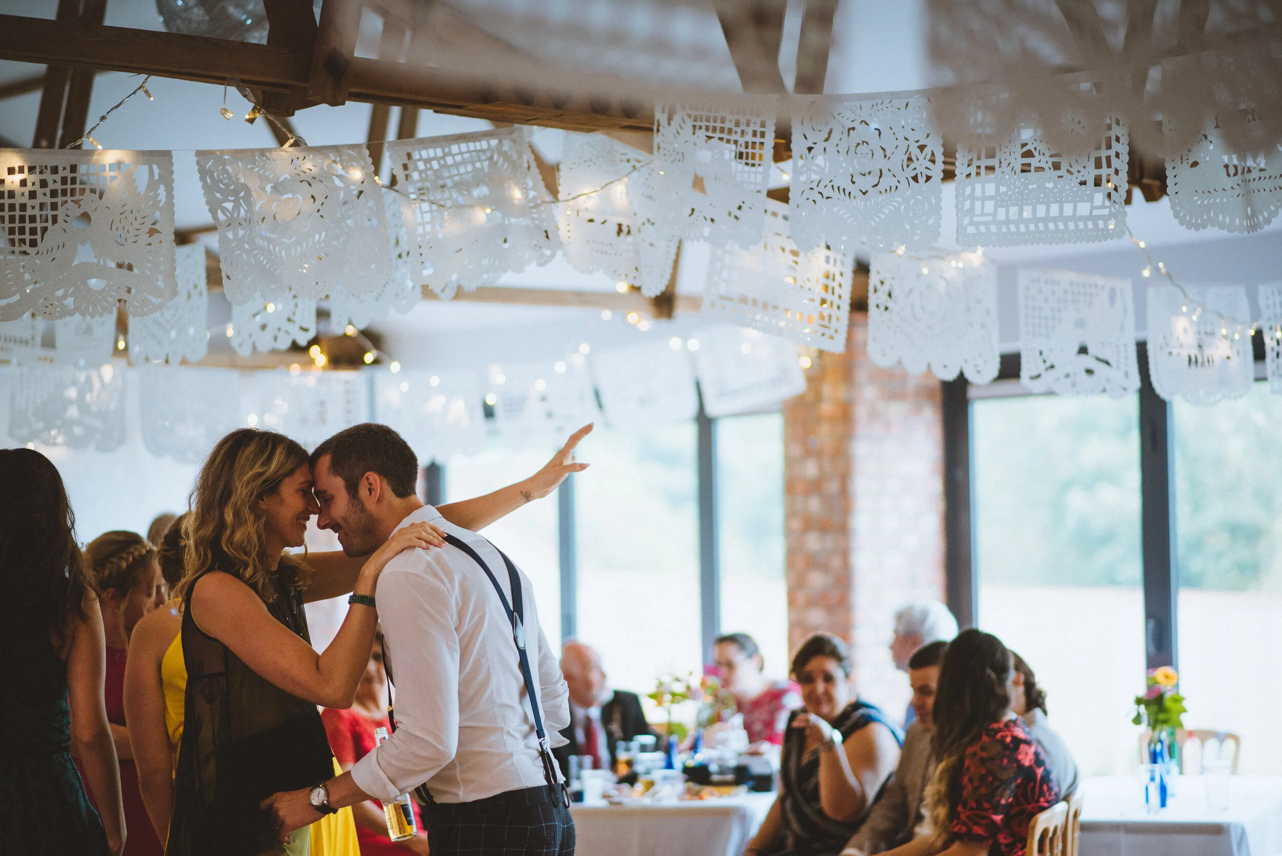 A couple dancing and smiling at a wedding reception with guests seated at tables in the background, decorated with paper banners and fairy lights.