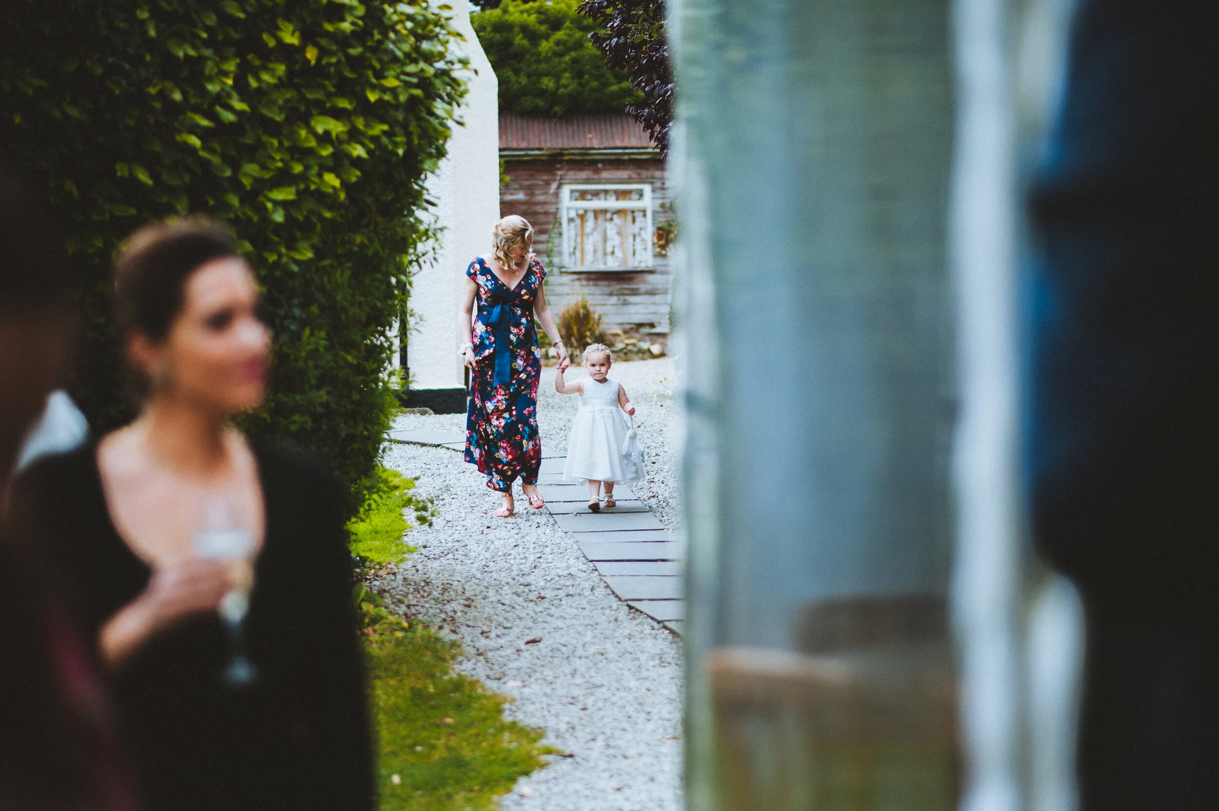 A woman wearing a floral dress walking hand-in-hand with a young girl in a white dress along a gravel pathway, with blurred figures of women on either side.