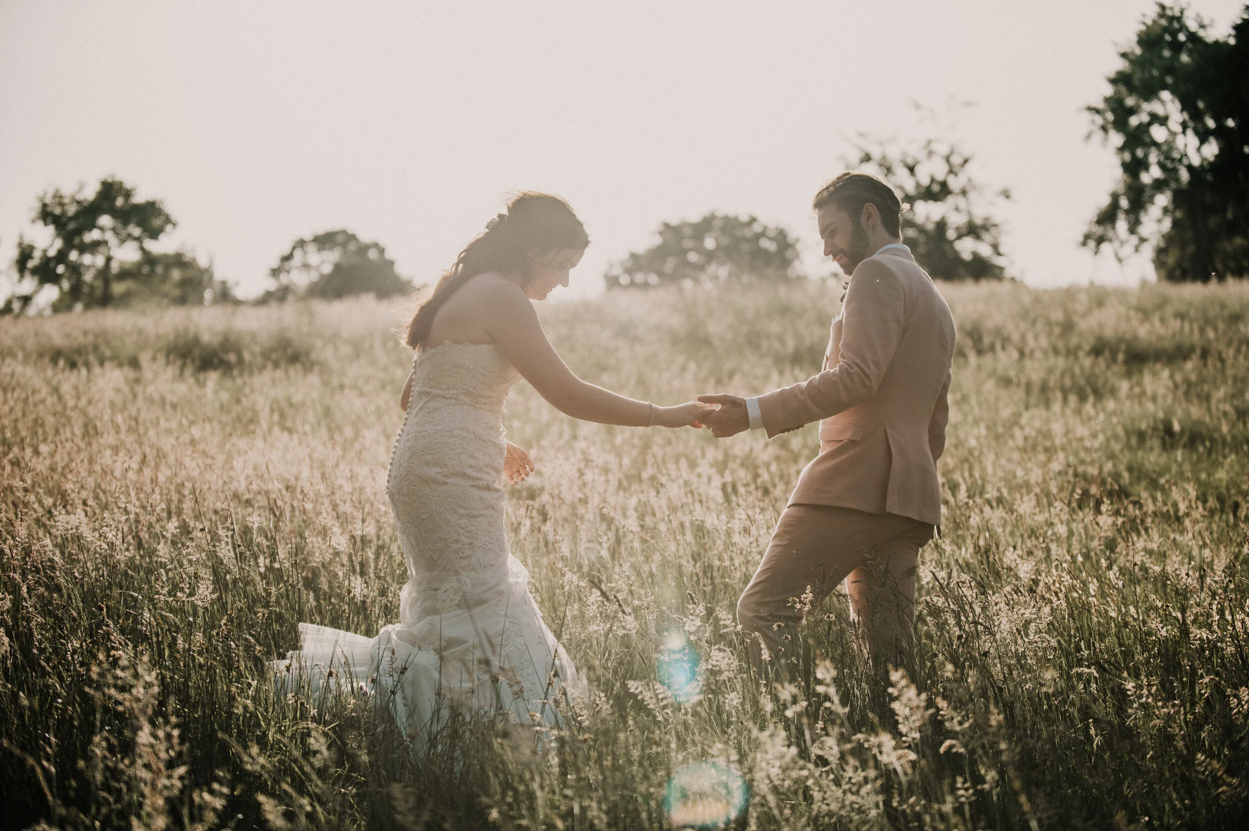 A bride and groom holding hands and dancing in a field during sunset