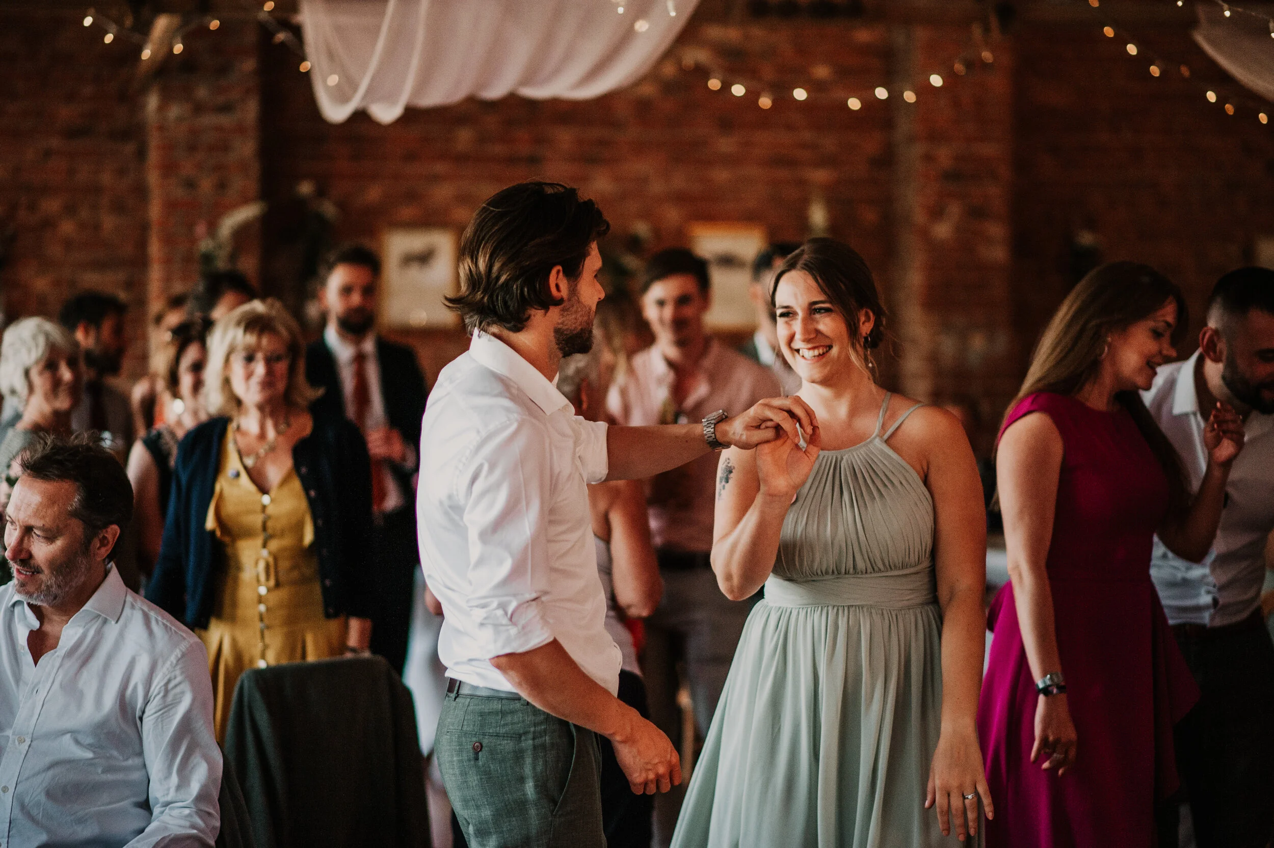 A couple dancing at a wedding reception, surrounded by guests in a rustic venue with brick walls and fairy lights.