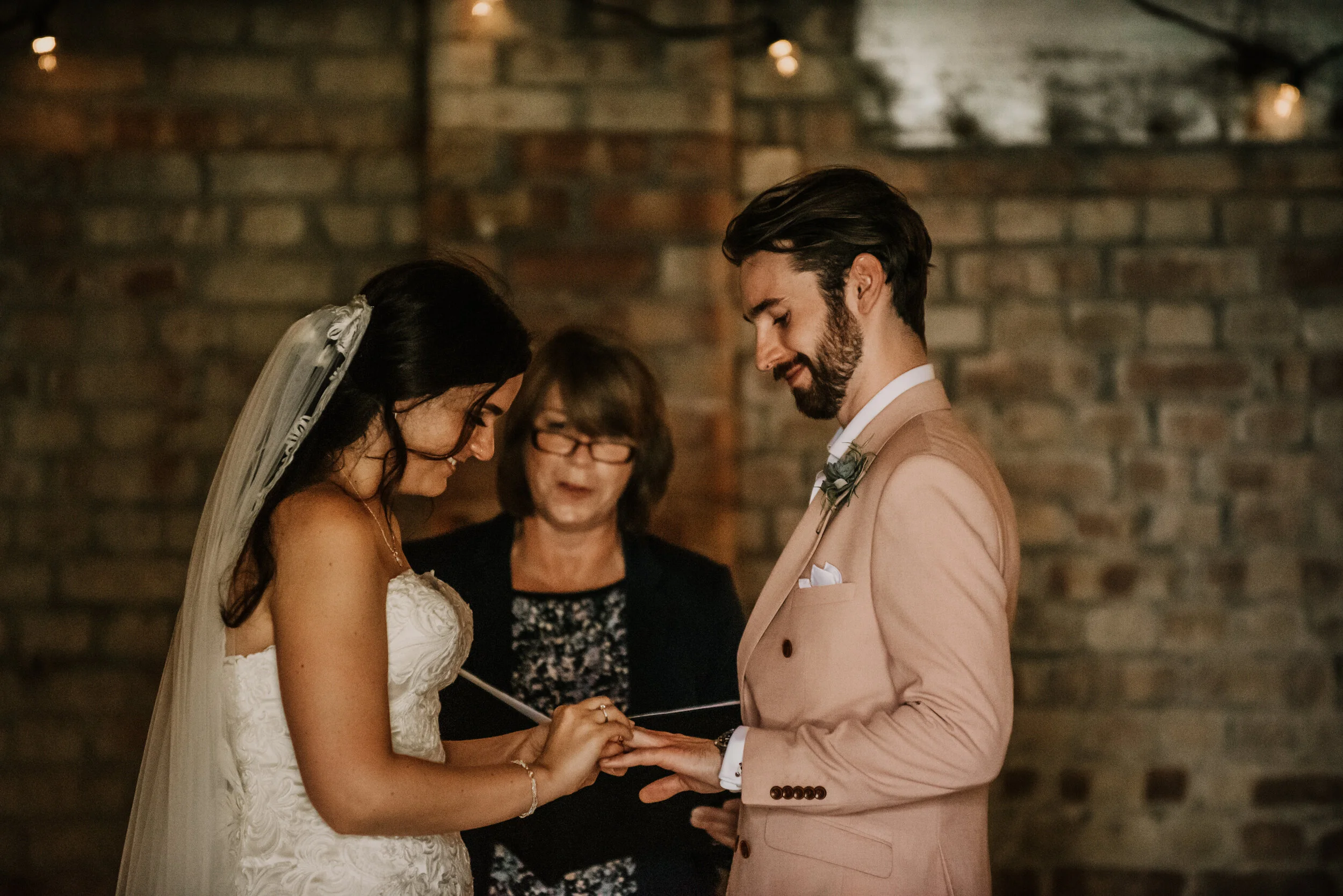 A bride and groom exchange rings during a wedding ceremony, with officiant standing behind them against a brick wall.