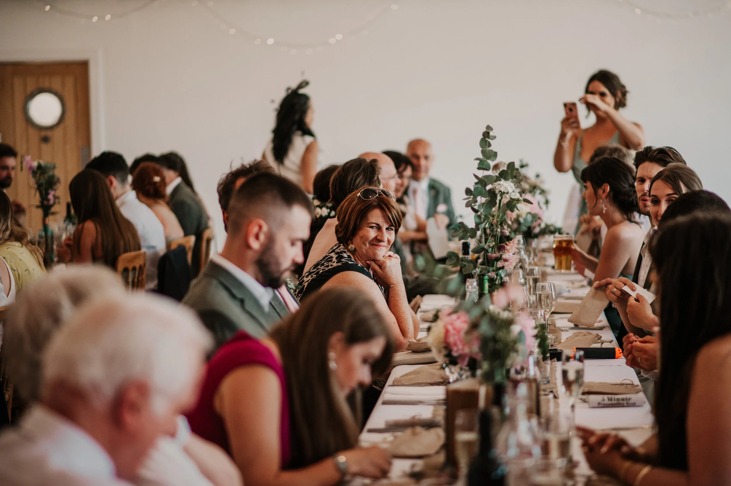People seated at a long table during a social event, possibly a wedding reception, with floral centerpieces and general festive decor.