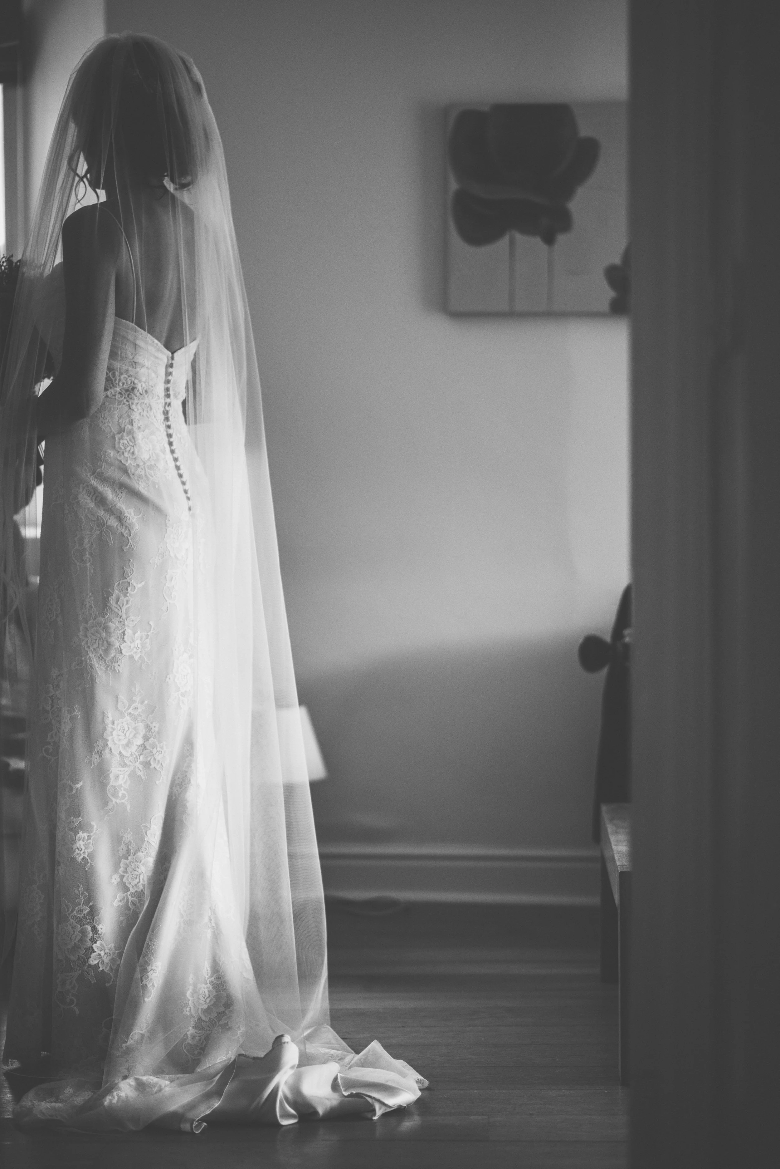 A woman in a wedding dress and veil standing inside a room with natural light coming through a window.