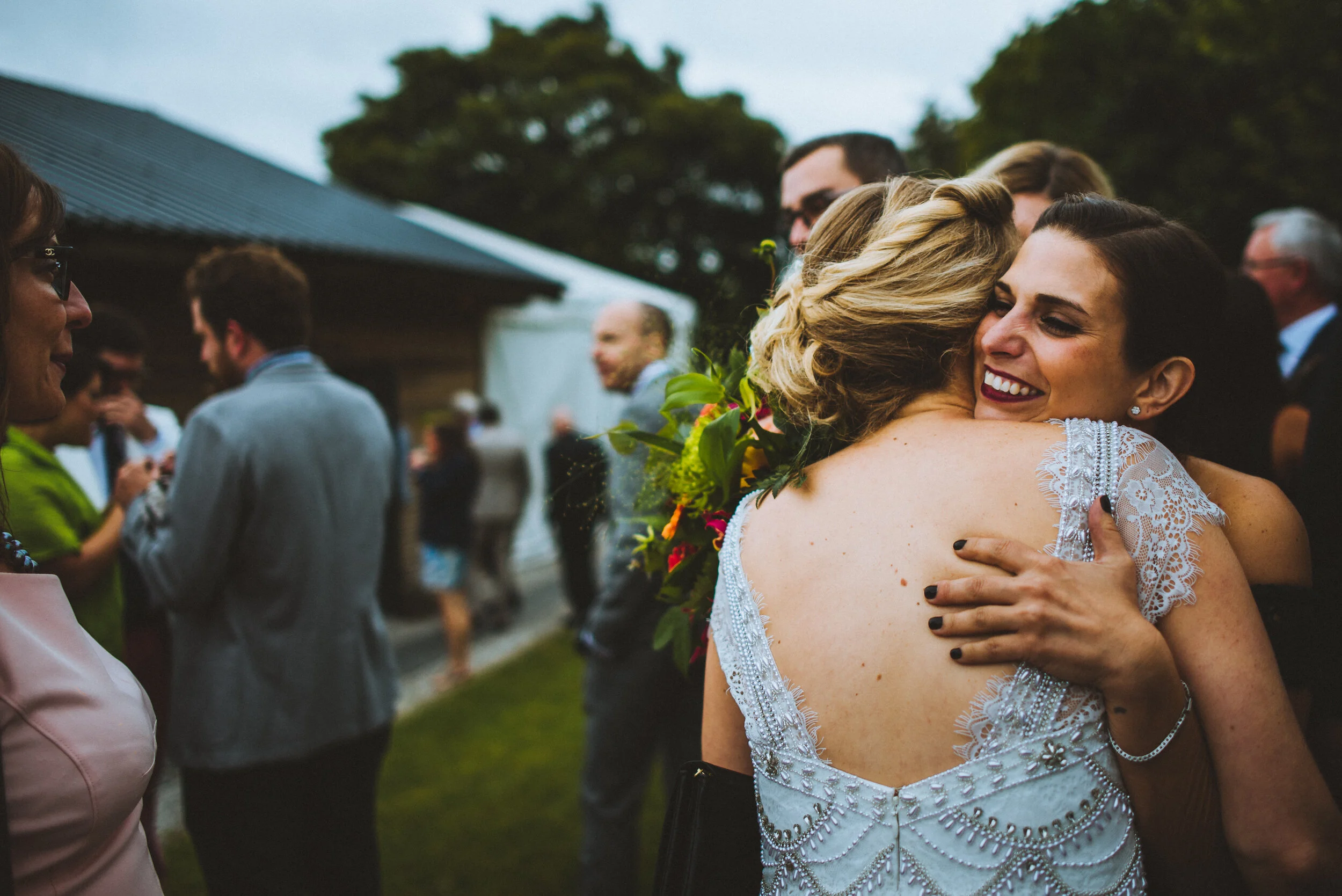 Two women hugging, one in a white dress with lace details, at an outdoor wedding. Several people are in the background, socializing near a building and trees.