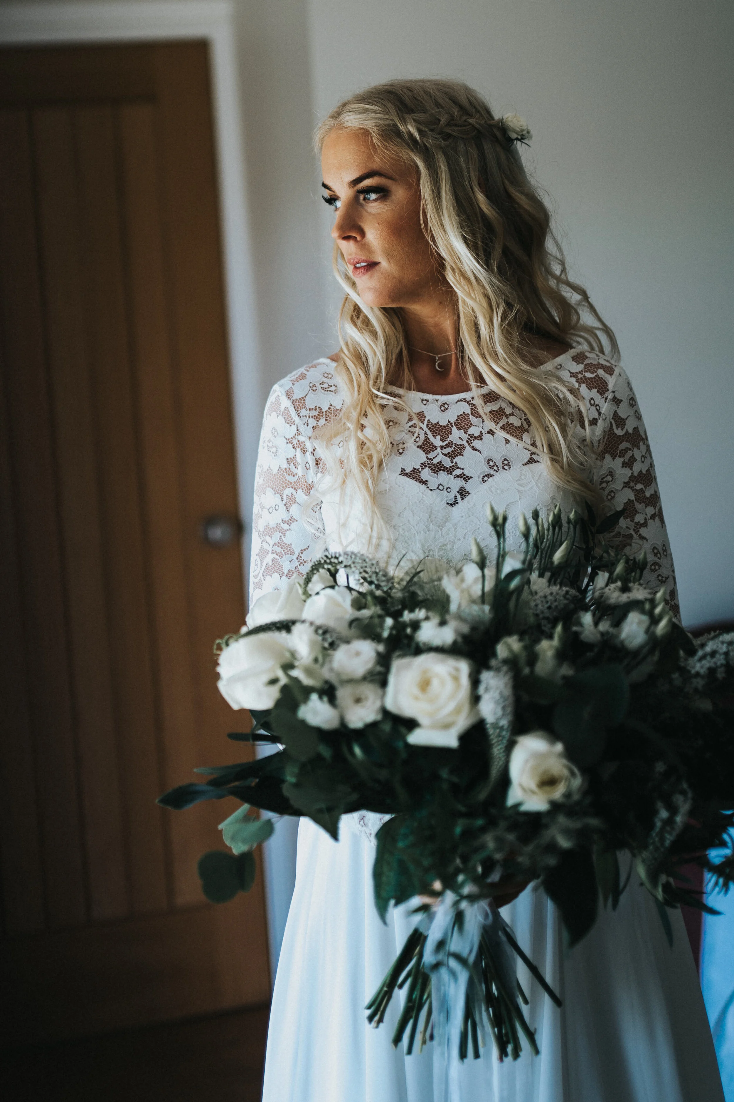 A woman with long blonde hair styled in loose curls, wearing a white lace dress, holding a bouquet of white flowers and greenery, standing indoors.