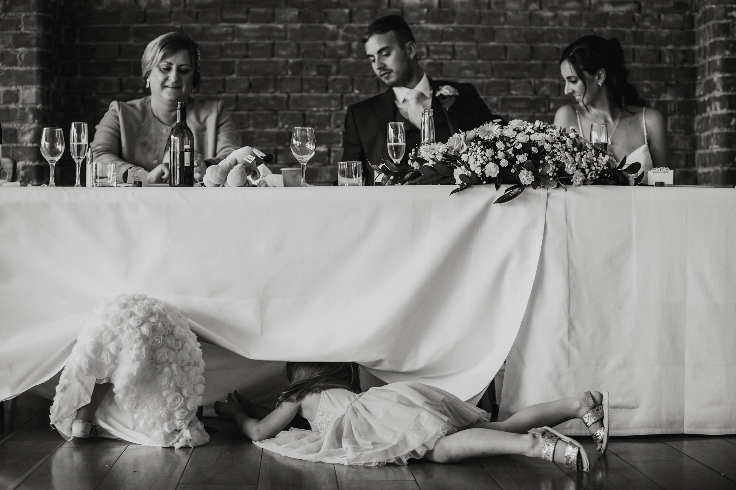 A little girl is crawling under a table at a wedding reception. The table is decorated with four glasses of wine, a bottle, a bunch of flowers, and a small cake.