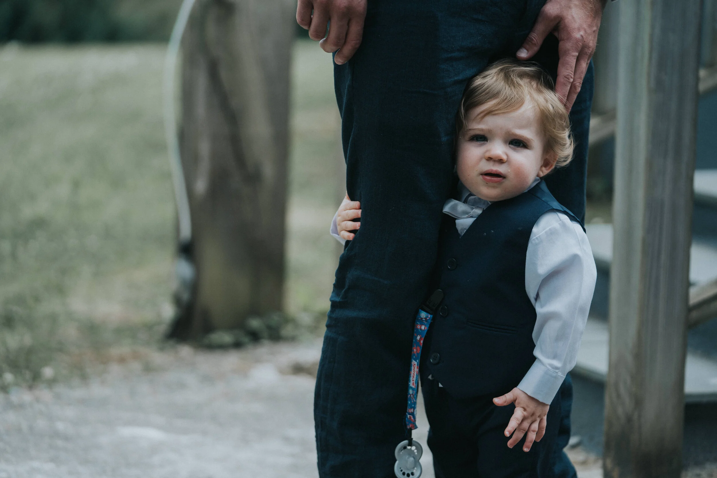 A young boy in formal attire, including a vest and bow tie, standing with his head against an adult's leg, holding onto the adult's hand with a curious or concerned expression on his face, outdoors near a wooden structure.