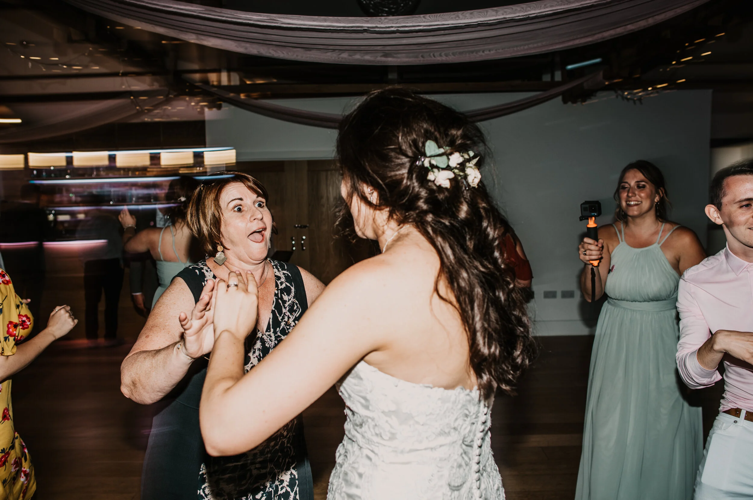 Two women, one in a black dress and the other in a white wedding gown, having fun dancing together at a wedding reception. Two other women are visible in the background, one holding a camera.