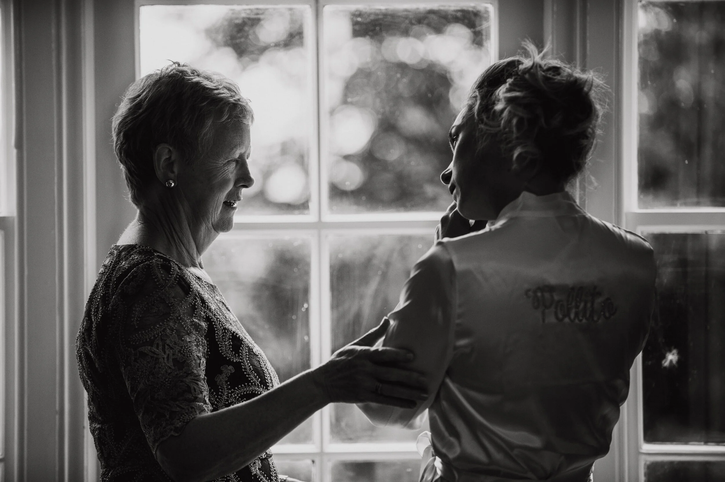 Two women facing each other and smiling, standing near a window, with sunlight illuminating their faces in a black and white photograph.