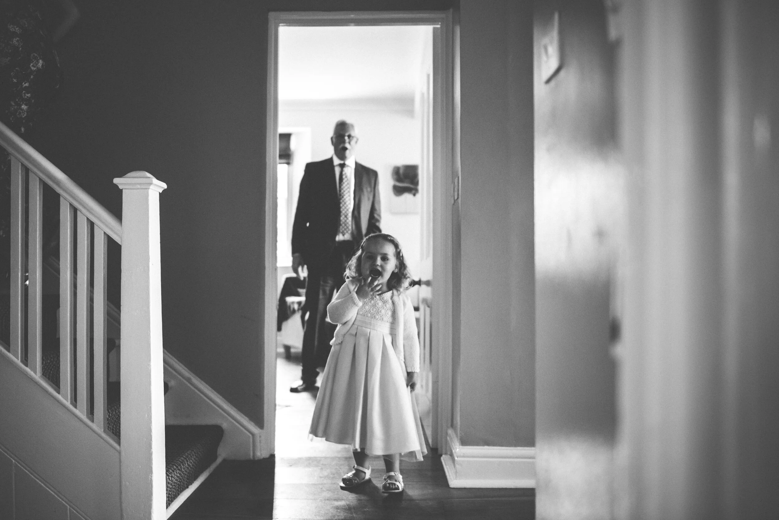 A young girl in a formal dress stands in a hallway, looking at the camera with her finger on her lips. Behind her, a man in a suit and tie stands in a doorway, slightly blurred in the background.