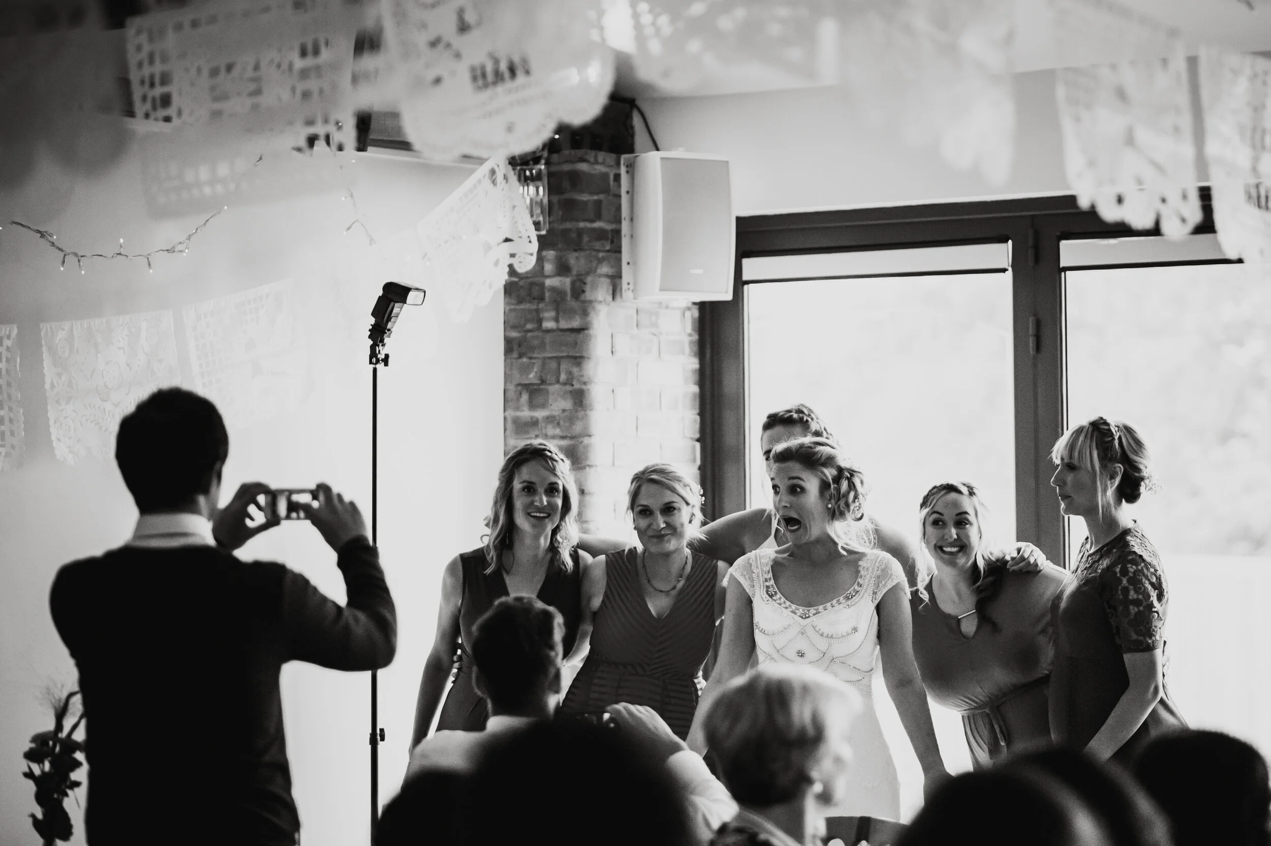 A group of women smiling and posing for a photo at a social gathering, taken indoors with large windows in the background.