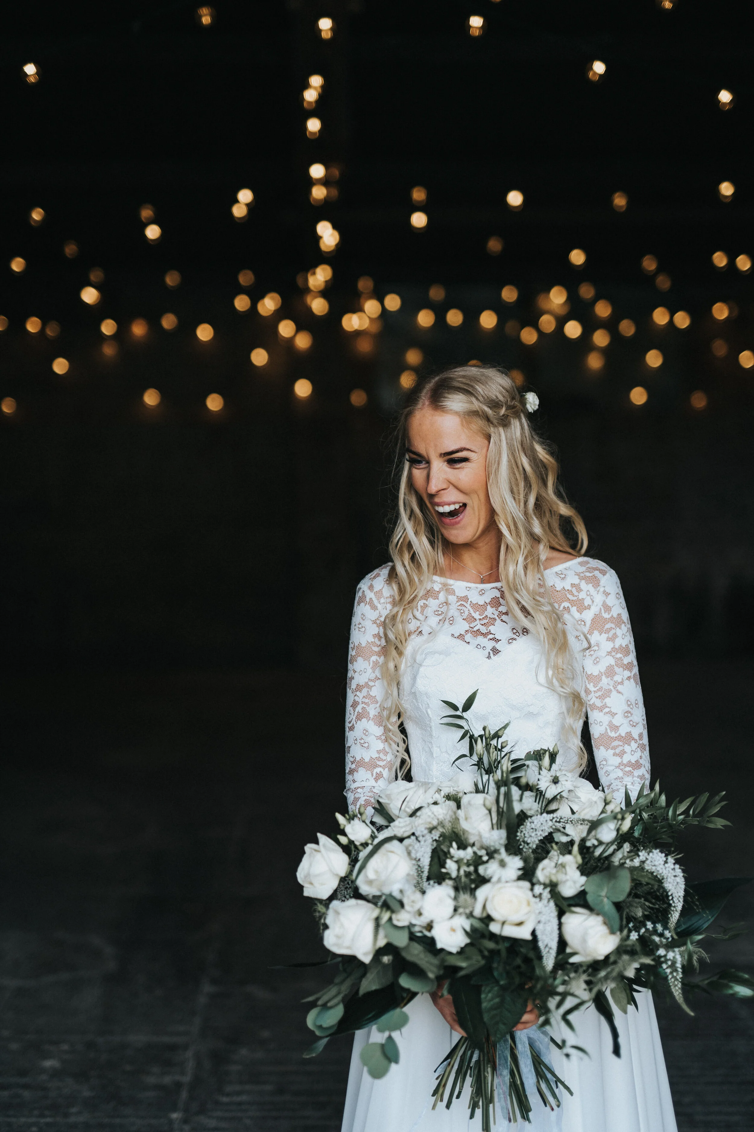 A bride in a white wedding dress with lace sleeves holding a large bouquet of white roses and greenery, smiling indoors with blurred warm string lights in the background.