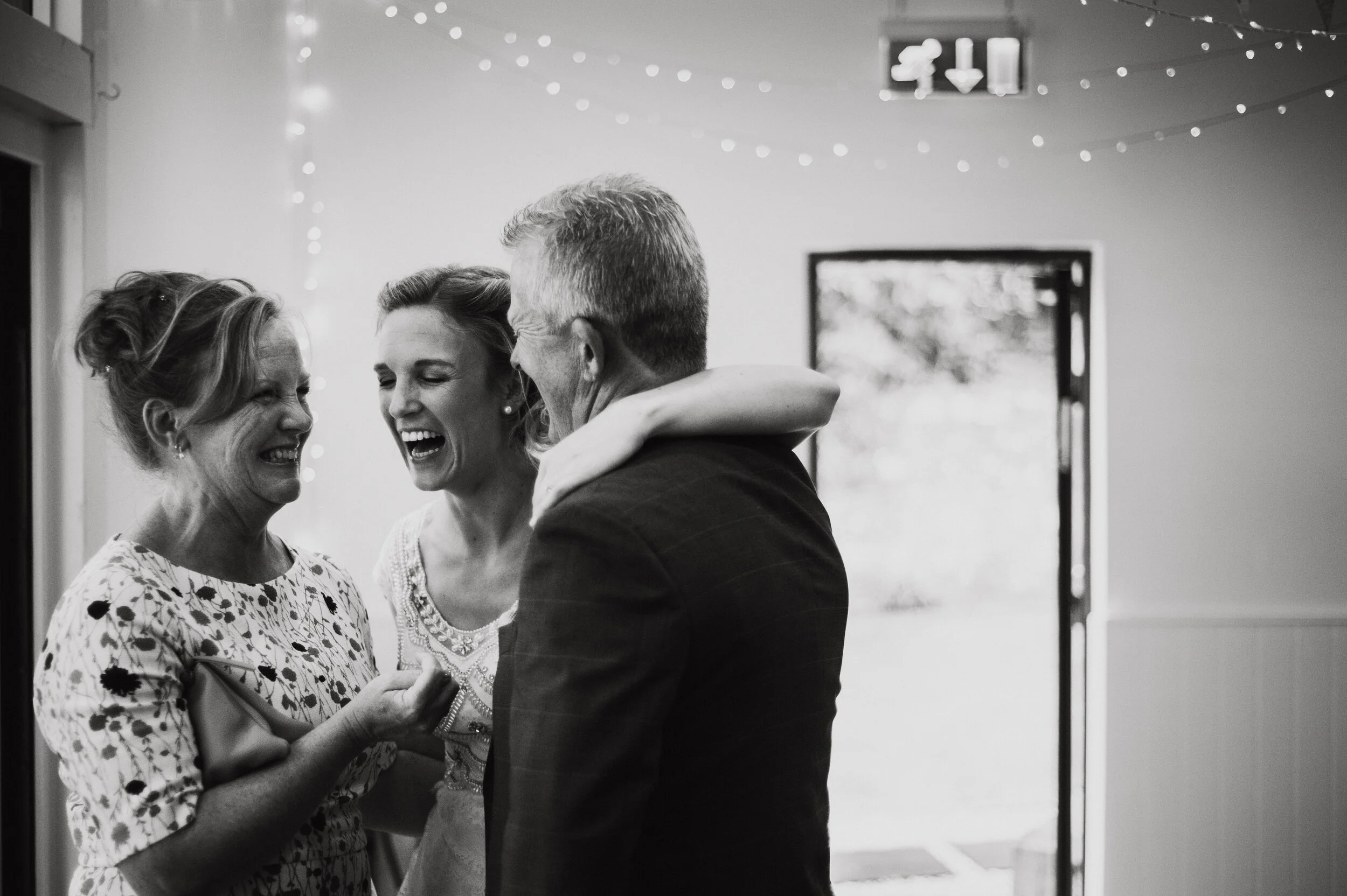 Three adults, two women and one man, are laughing and smiling inside a decorated room. The woman on the left has her hair tied up and is wearing a patterned dress. The woman in the middle has short hair and is wearing a lace top. The man on the right