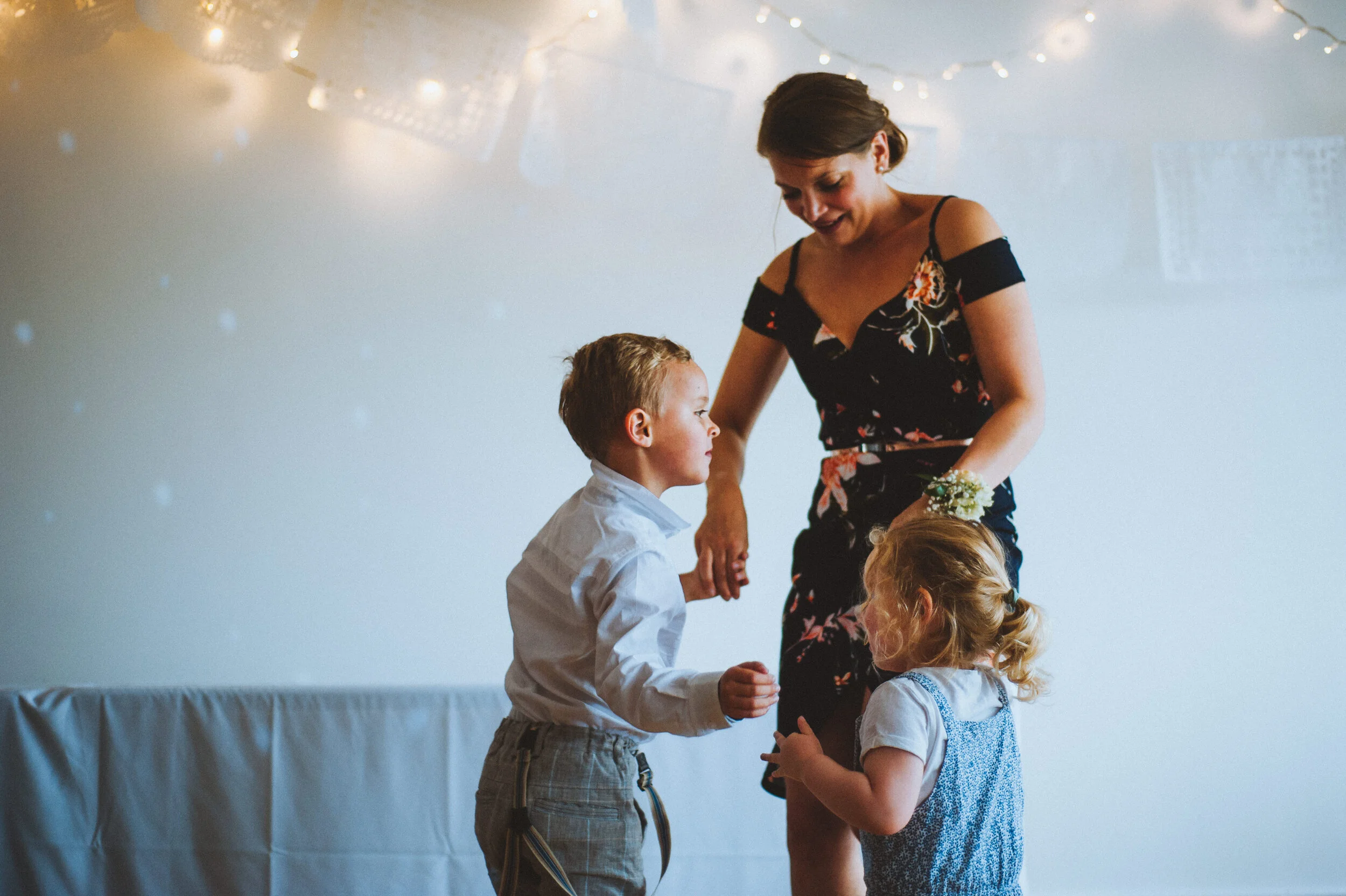 A woman with dark hair in a floral dress holding hands and talking to two children, a boy and a girl, at what appears to be a celebration or party, with string lights in the background.