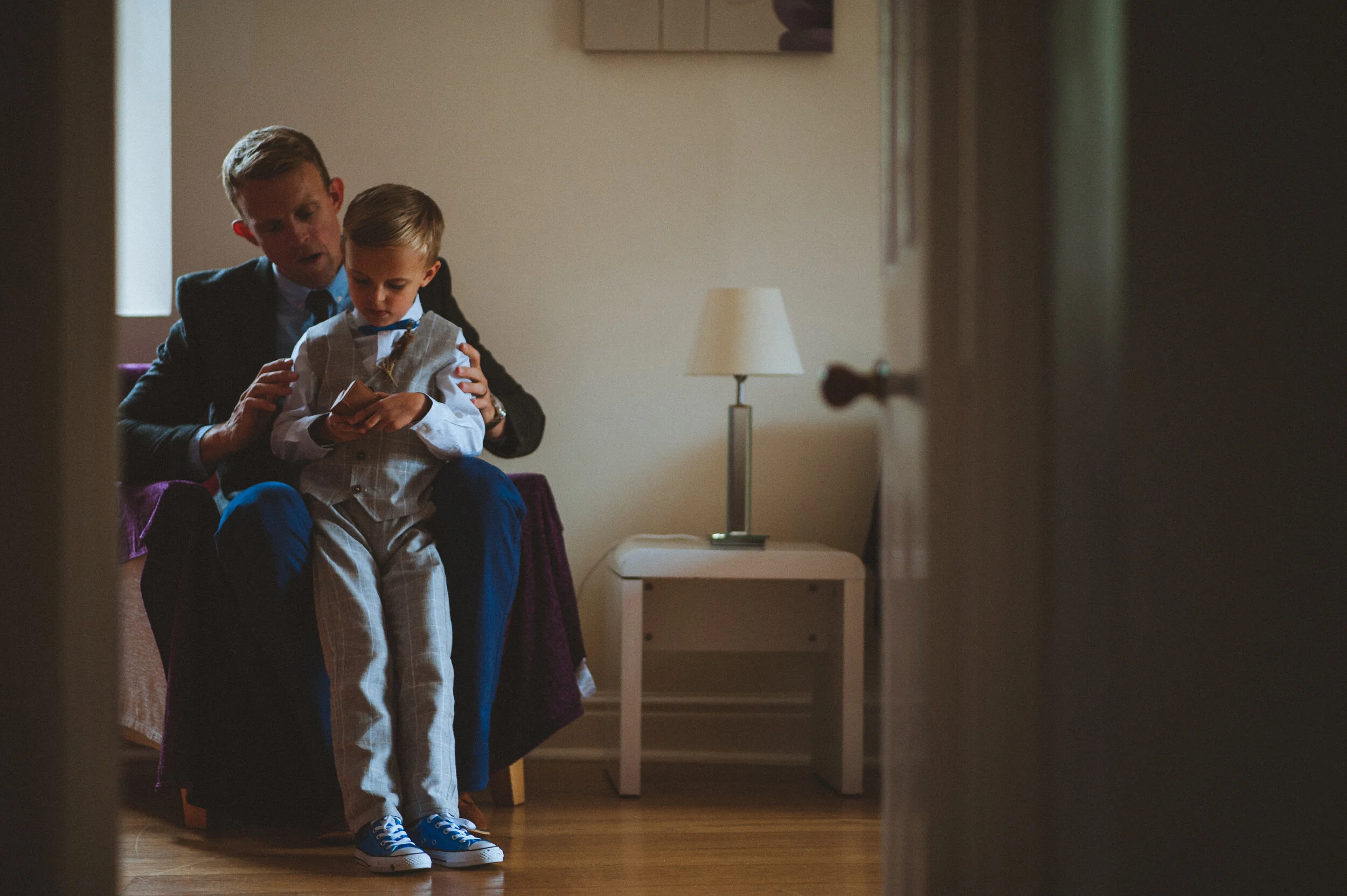 A man in a suit sits on a purple couch with a young boy in a suit sitting on his lap, both looking at a phone. The scene is in a cozy room with a white table and a lamp on it, visible in the background.
