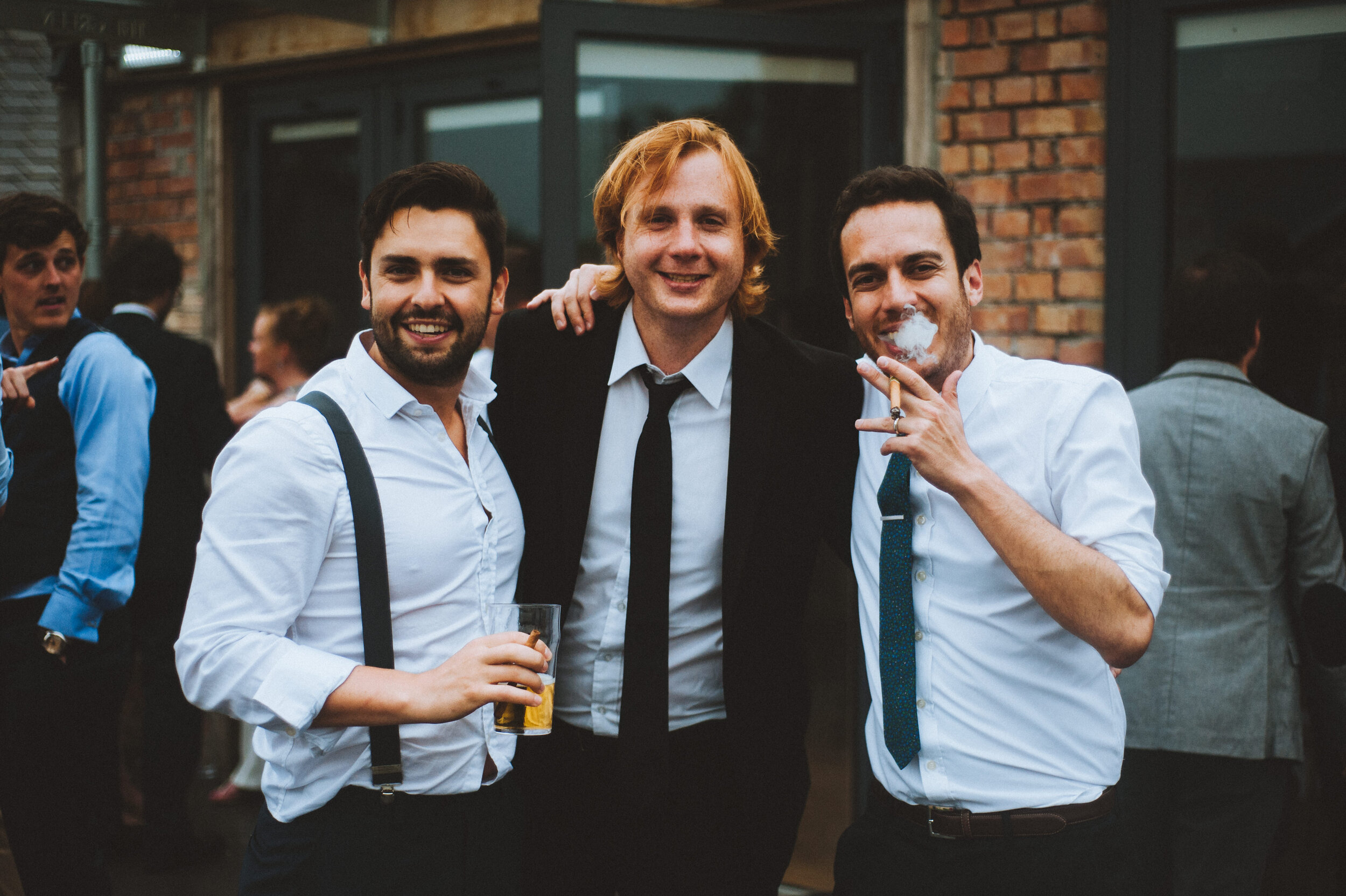 Three men dressed in business casual attire posing together at a social gathering outside a brick building, smiling and holding drinks, with other people in the background.
