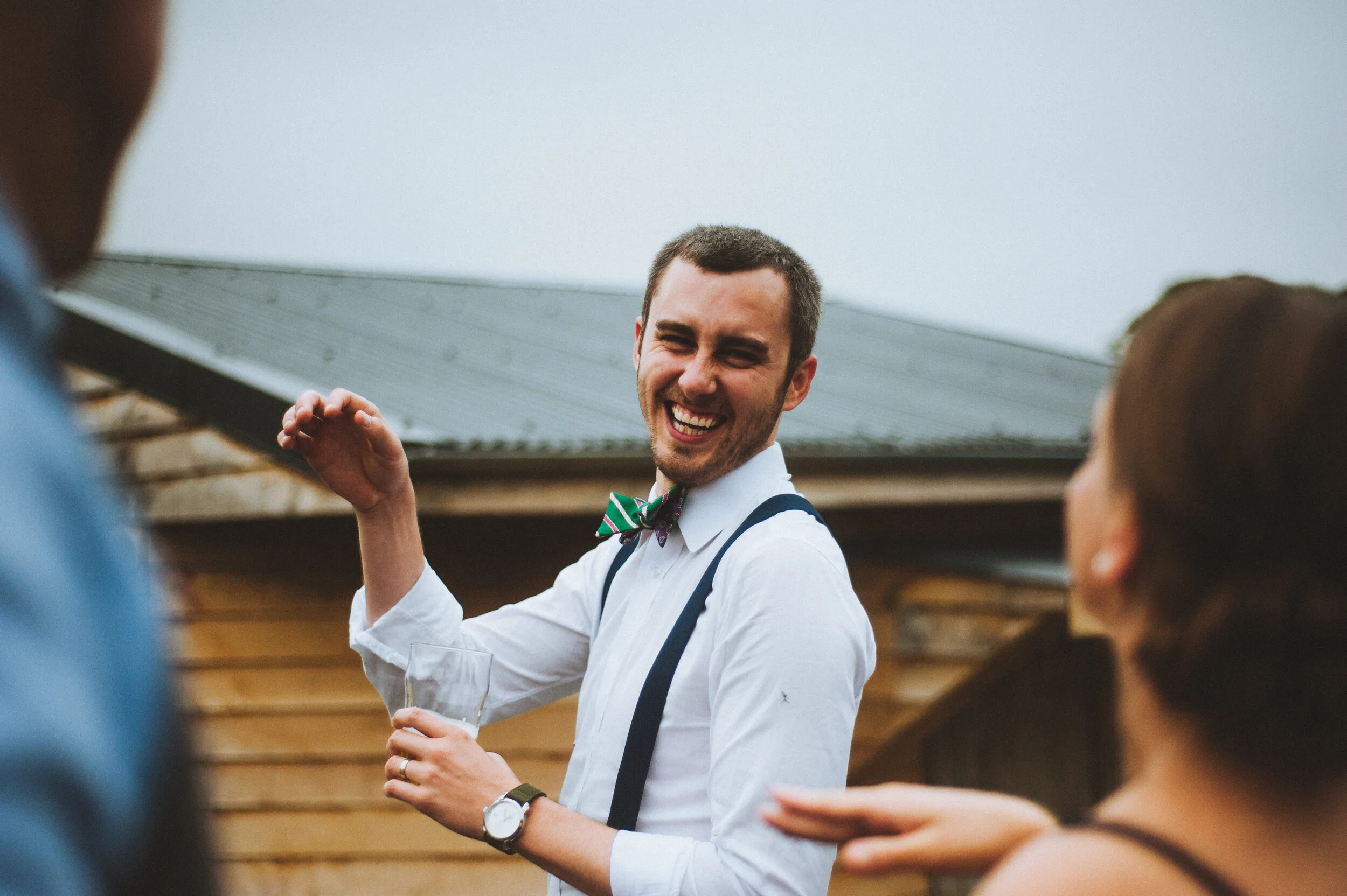 Man with a bowtie laughing and holding a drink, talking to two women outdoors near a wooden shed.