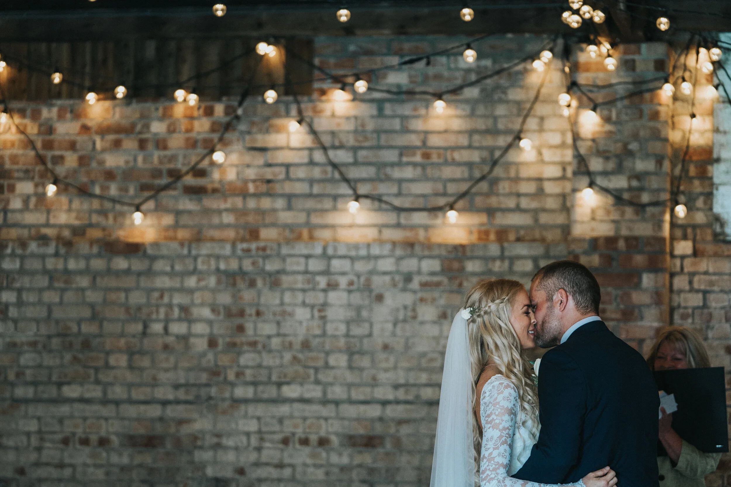 A bride and groom sharing a kiss during their wedding ceremony, with fairy lights hanging on a brick wall in the background.