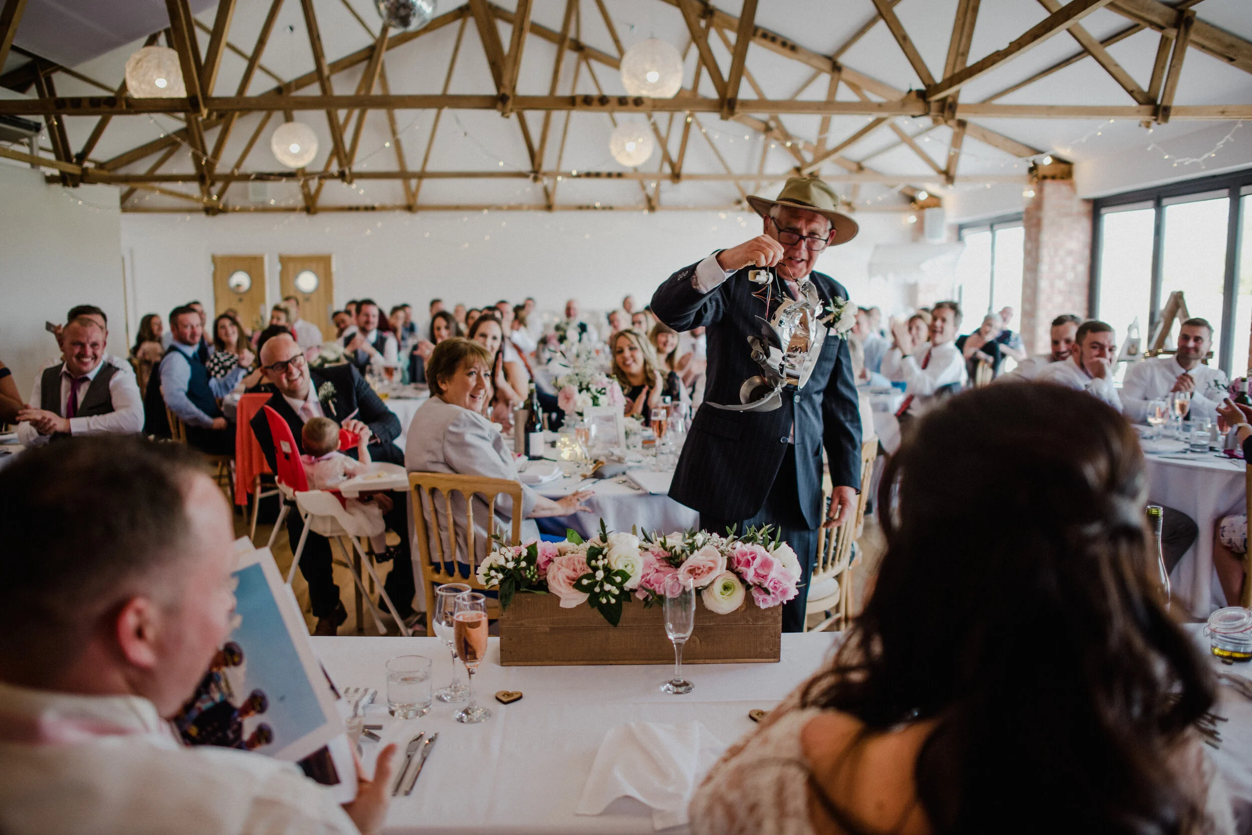 A man in a dark suit and hat giving a toast at a wedding reception, surrounded by seated guests in a decorated banquet hall.