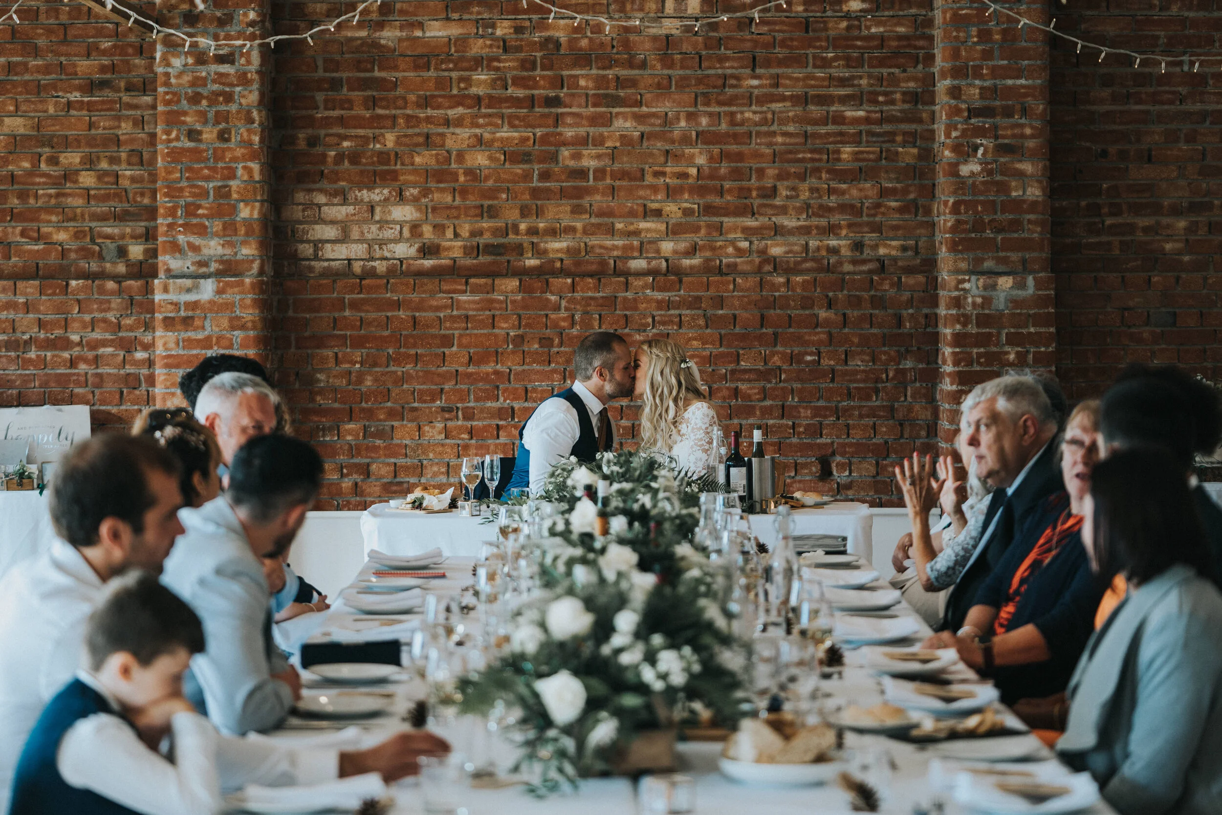 A wedding reception with the bride and groom sharing a kiss at their head table, surrounded by seated guests, decorated with a white tablecloth and floral centerpiece against a brick wall.
