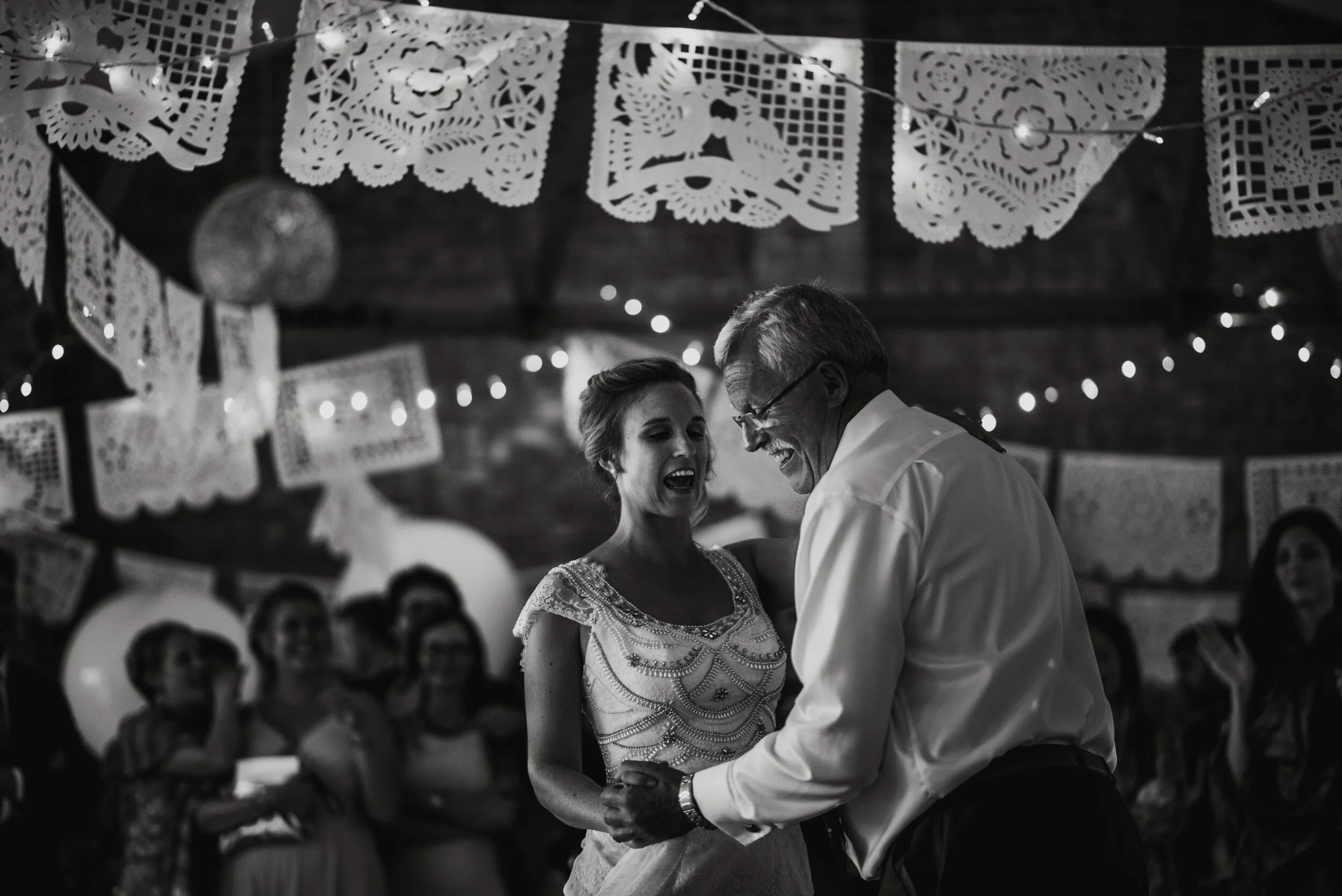 A black and white photo of a bride and an older man dancing together at a celebration or wedding reception. The background features decorative paper banners and string lights, with a group of people watching and smiling.