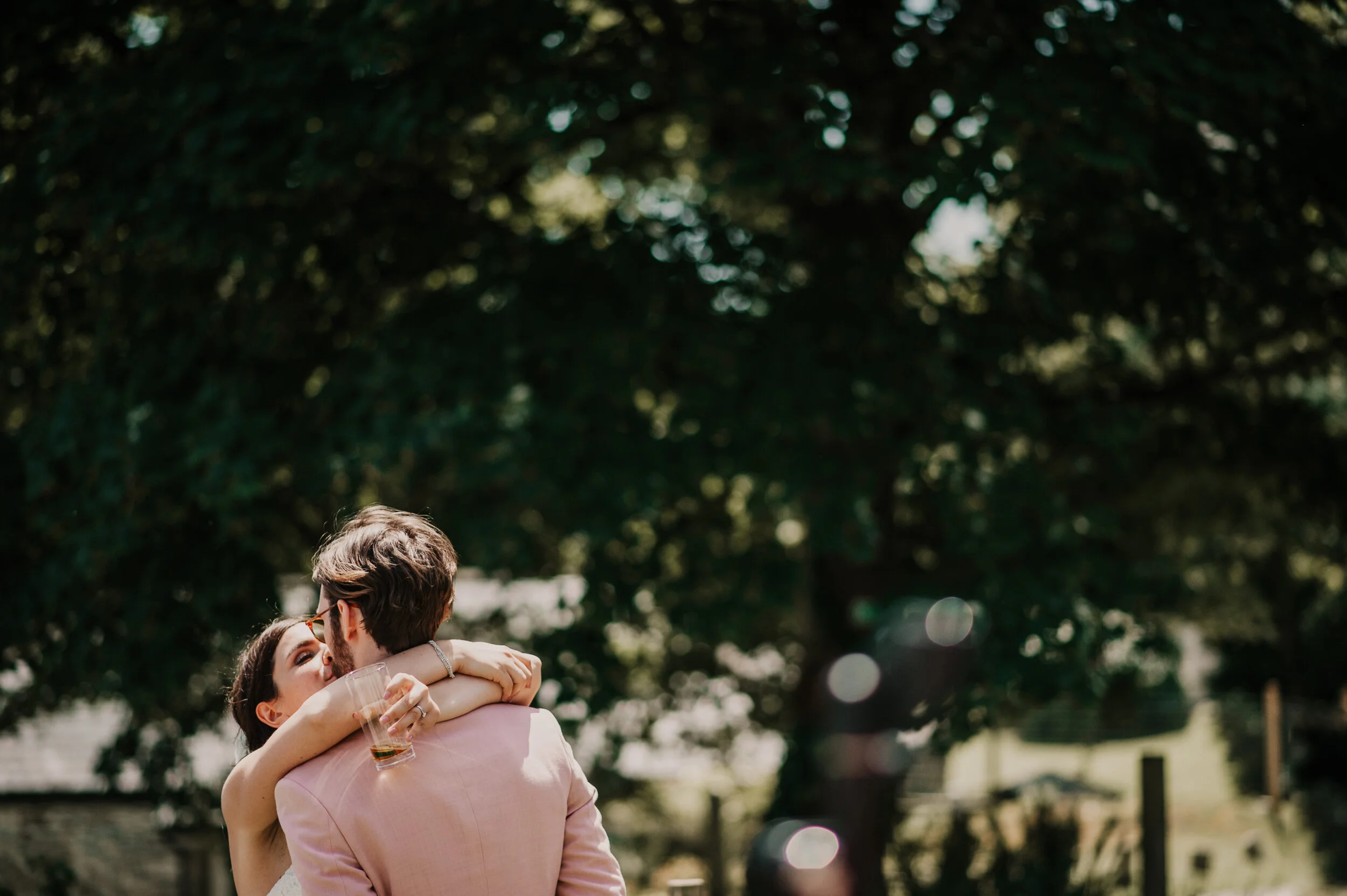 A couple sharing a romantic moment outdoors, with the woman hugging the man and holding a glass, in a park with trees in the background.