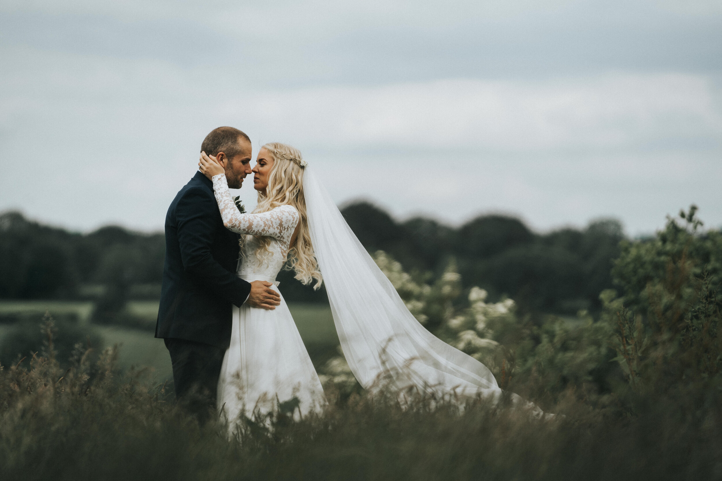 A bride and groom in wedding attire embrace outdoors in a field, with a cloudy sky and distant trees in the background.