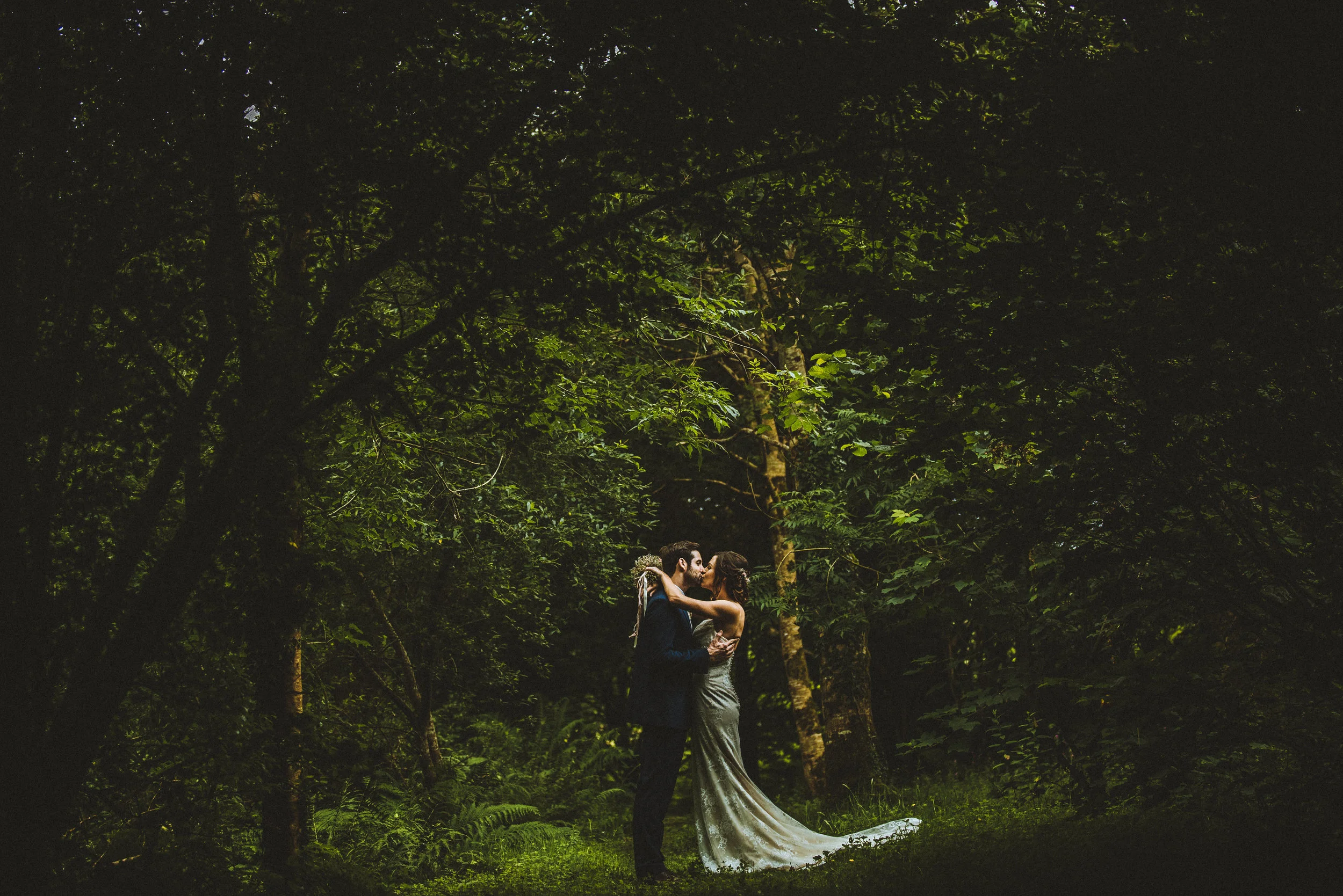 A bride and groom share a kiss in a forest at night. The bride wears a long, elegant gown, and the groom is dressed in a dark suit. They are surrounded by lush green trees and ferns.
