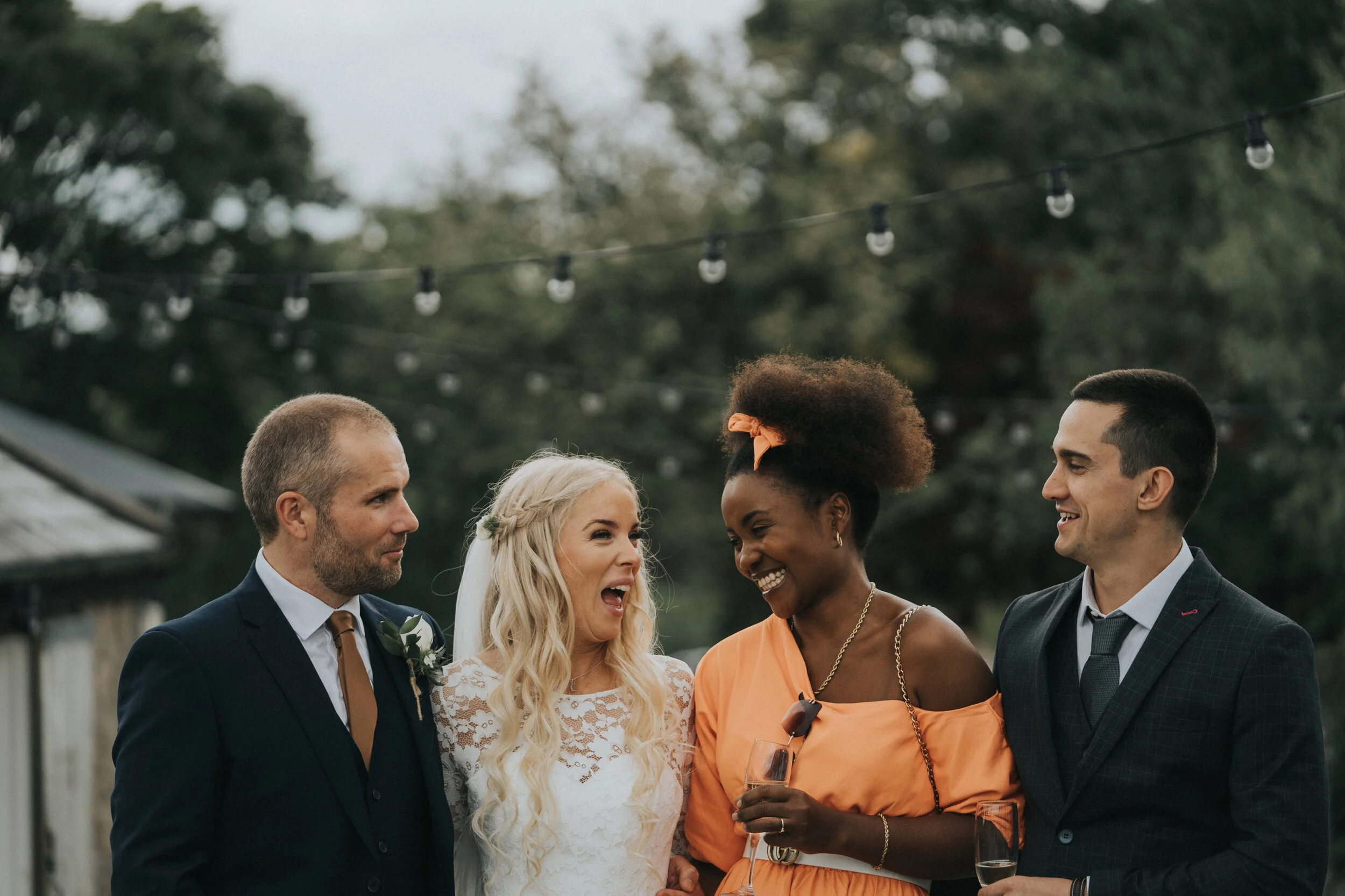 Group of four people, two men and two women, at an outdoor wedding reception, smiling and chatting, holding glasses of wine, with string lights and trees in the background.