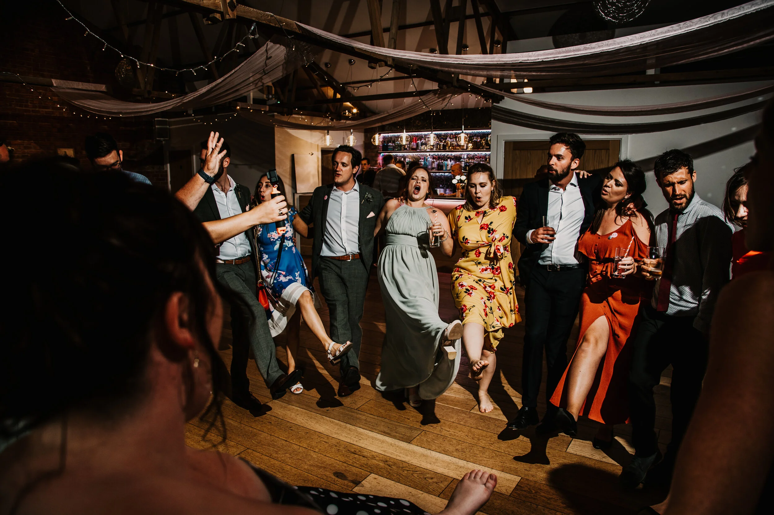 Group of people dancing and enjoying themselves at a party or celebration, with some holding drinks and others singing along, under decorative drapes and string lights.