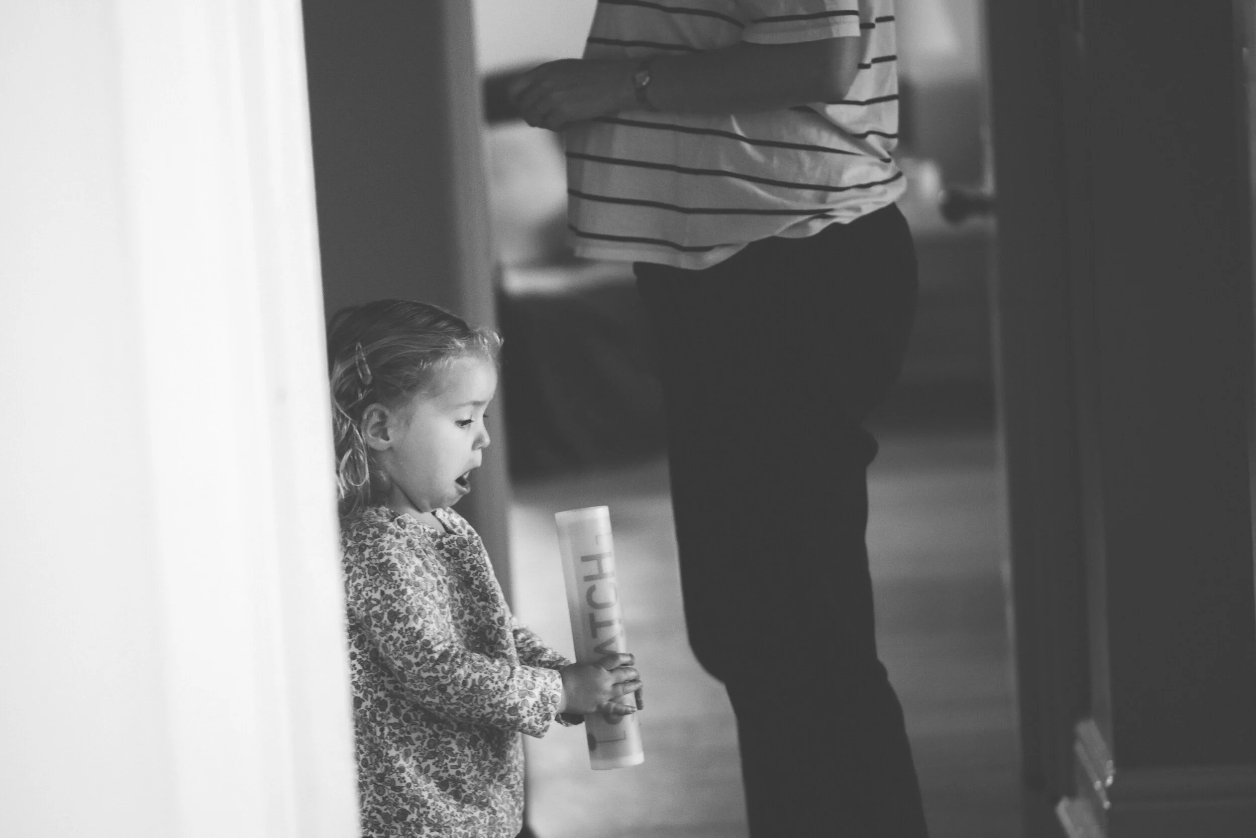 A young girl with curly hair holding a cylindrical object, standing against a wall, looking downward with her mouth open, while an adult woman in a striped shirt and dark pants stands nearby with her arms crossed.