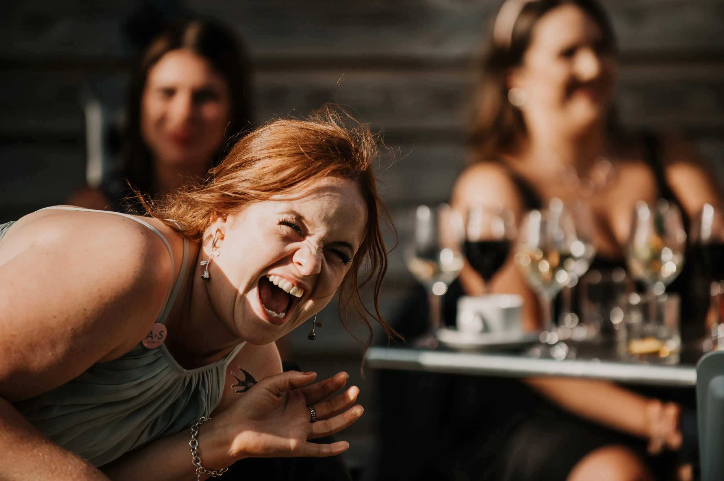 A woman laughing enthusiastically at a table with drinks, with other women blurred in the background.