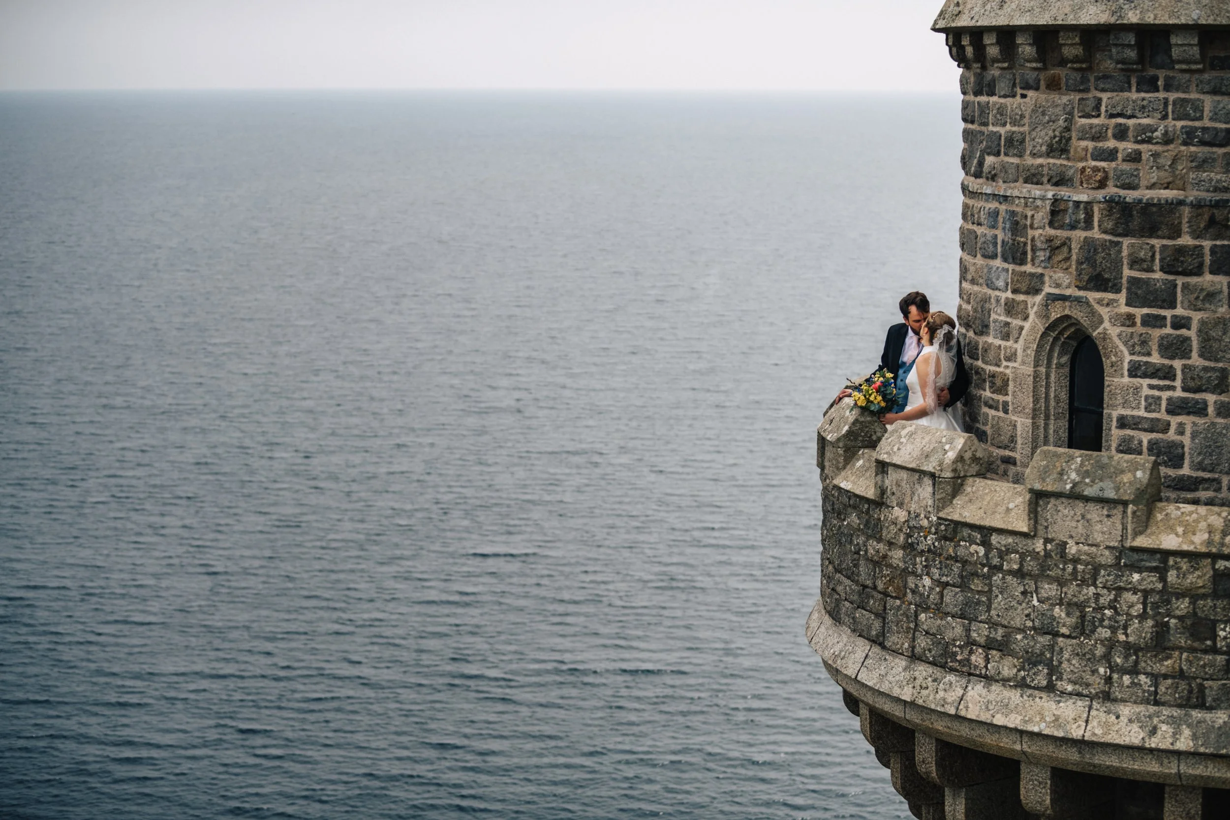A bride and groom sharing a kiss on a stone balcony of a castle overlooking the ocean.