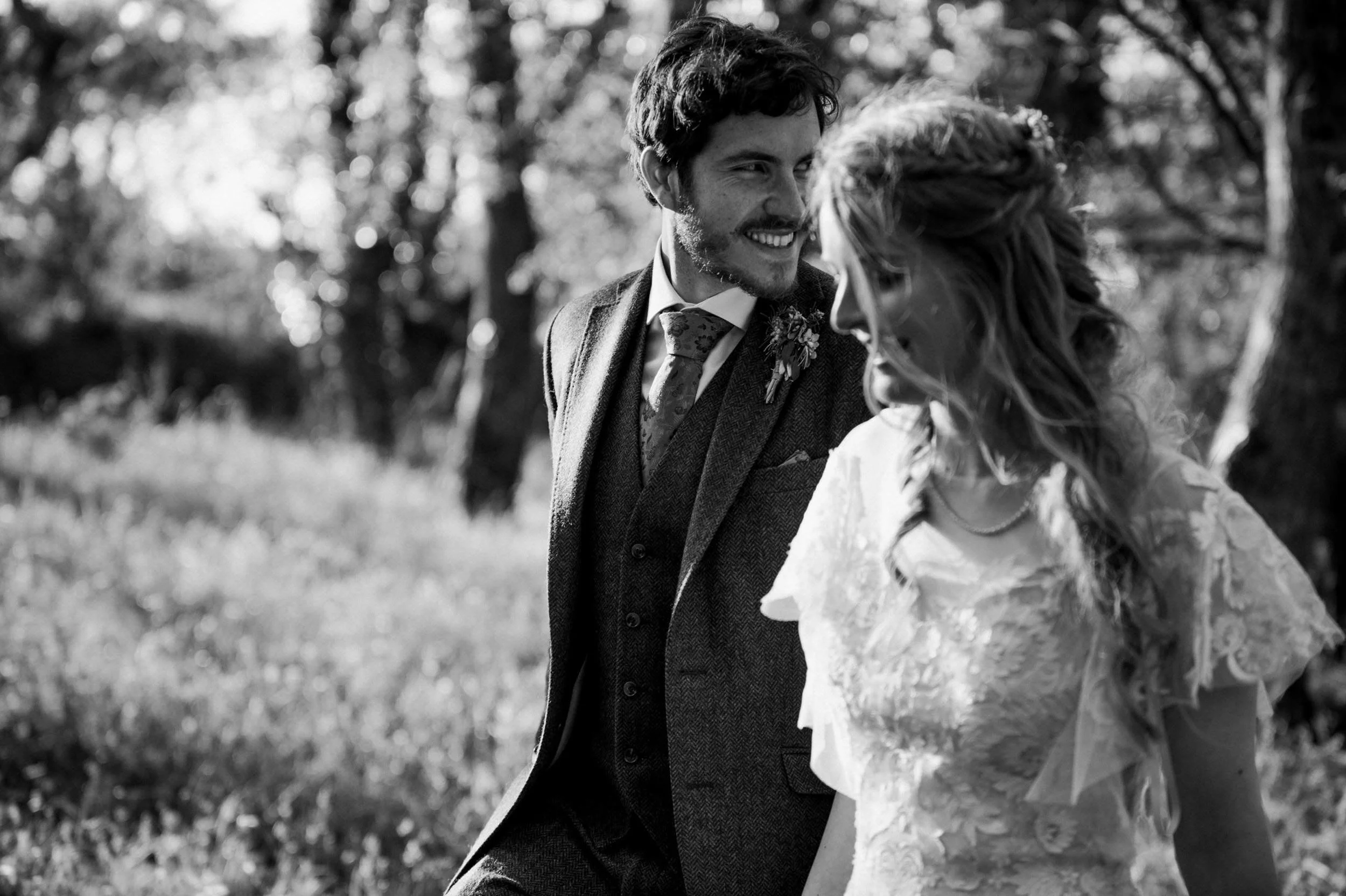 A black-and-white photo of a man and woman outdoors, smiling and looking at each other in a natural setting with trees and grass.