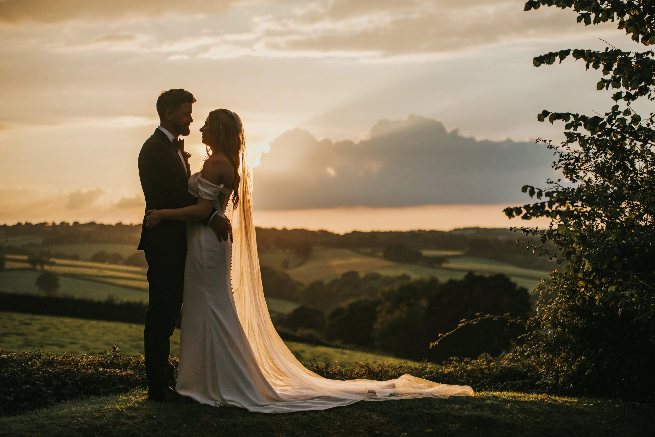 A bride and groom standing close together outdoors during sunset, with rolling hills and cloudy sky in the background.