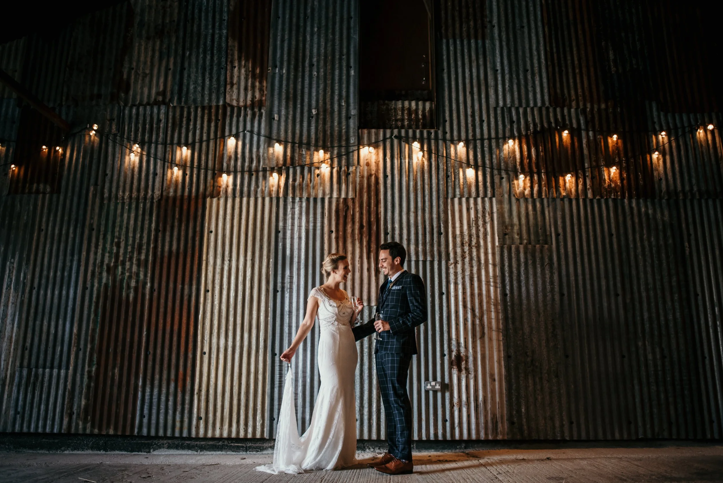 A bride and groom stand together in front of a corrugated metal wall decorated with string lights, sharing a moment at their wedding.