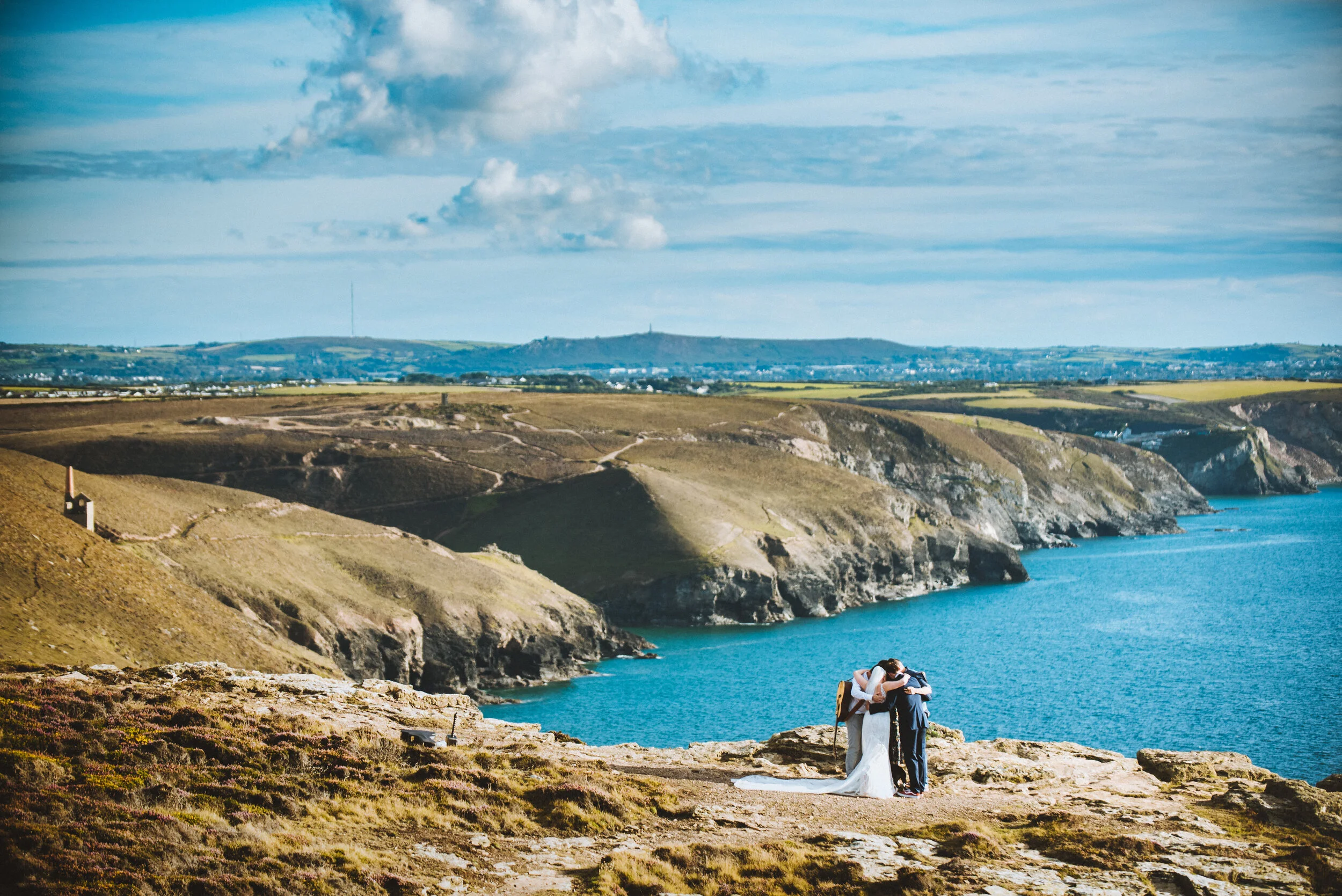 A bride and groom hugging on a rocky cliff overlooking the ocean with rolling hills in the background.