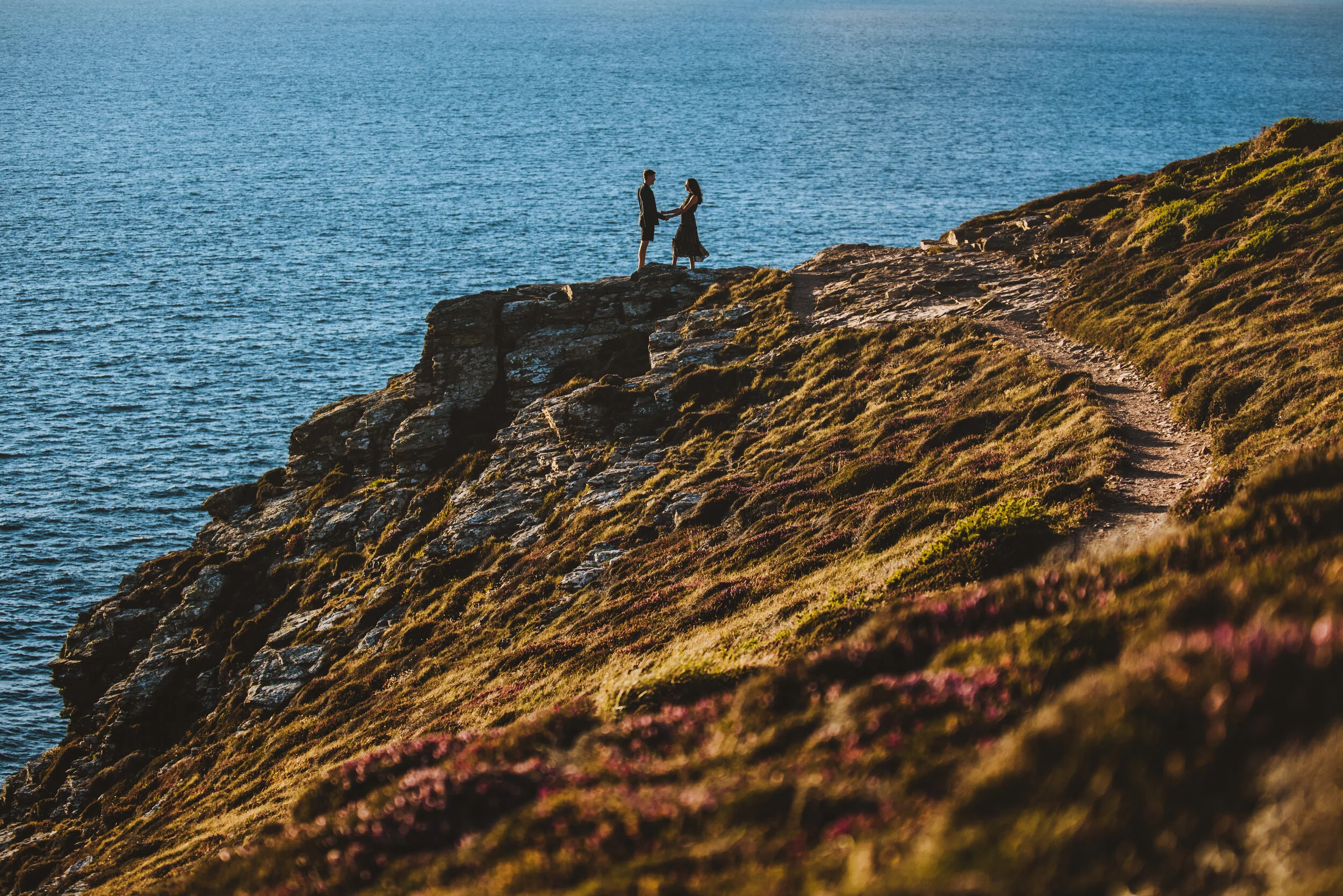 A couple holding hands stands on a rocky cliff overlooking the ocean during sunset.