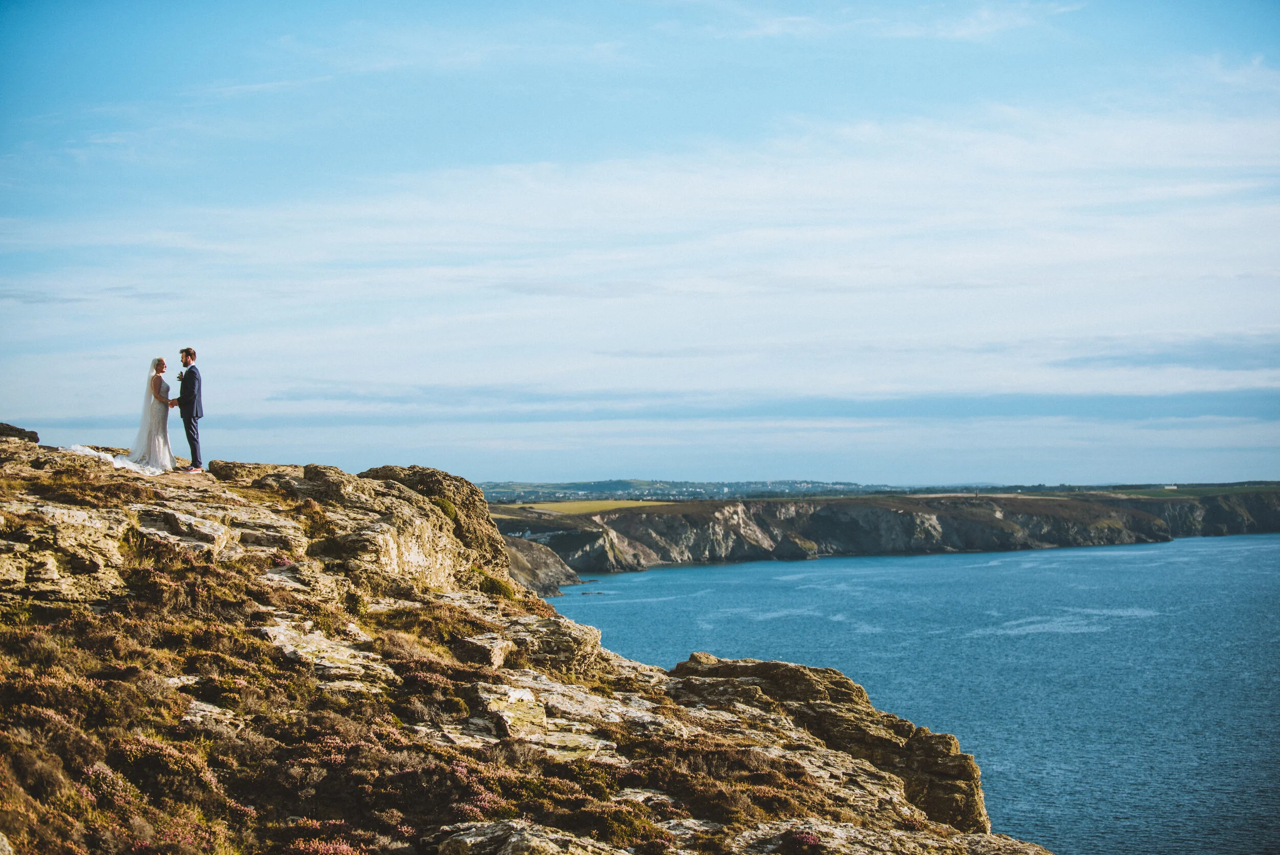 A bride and groom holding hands on a rocky cliff overlooking the ocean during their wedding photoshoot.