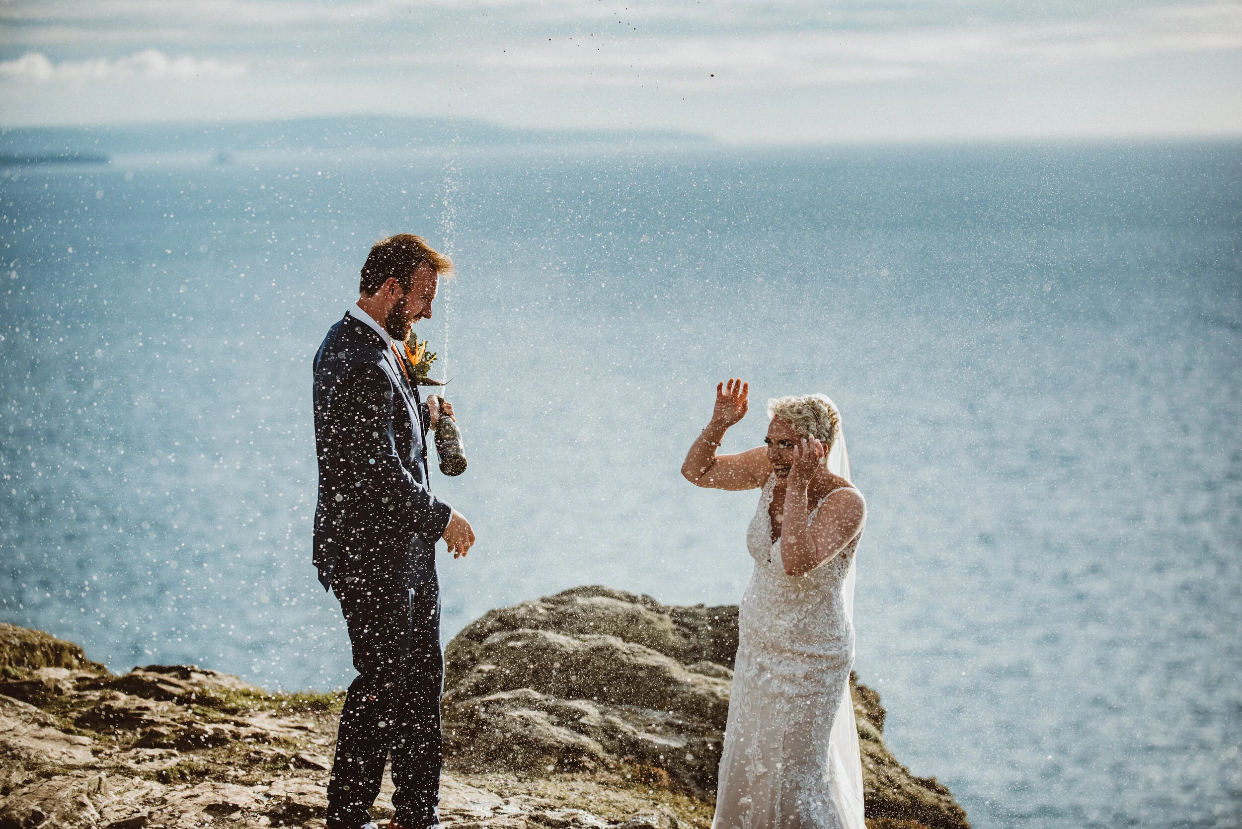 A bride and groom having a wedding ceremony on a rocky beach, with the bride laughing as water splashes around her and the groom holding a bottle.