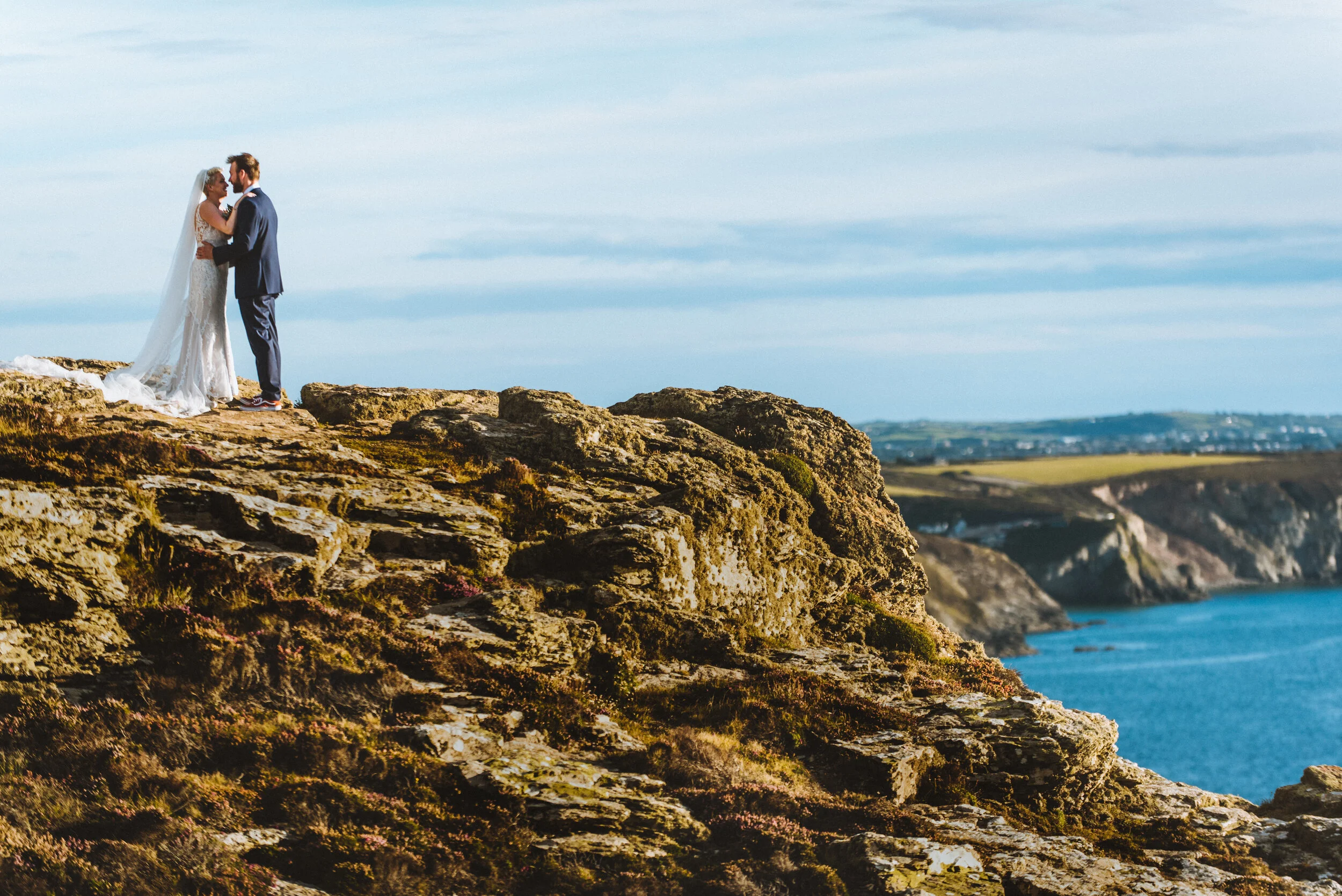 A bride and groom standing on a rocky cliff facing each other, with a view of the ocean and coastline in the background, during their wedding ceremony.