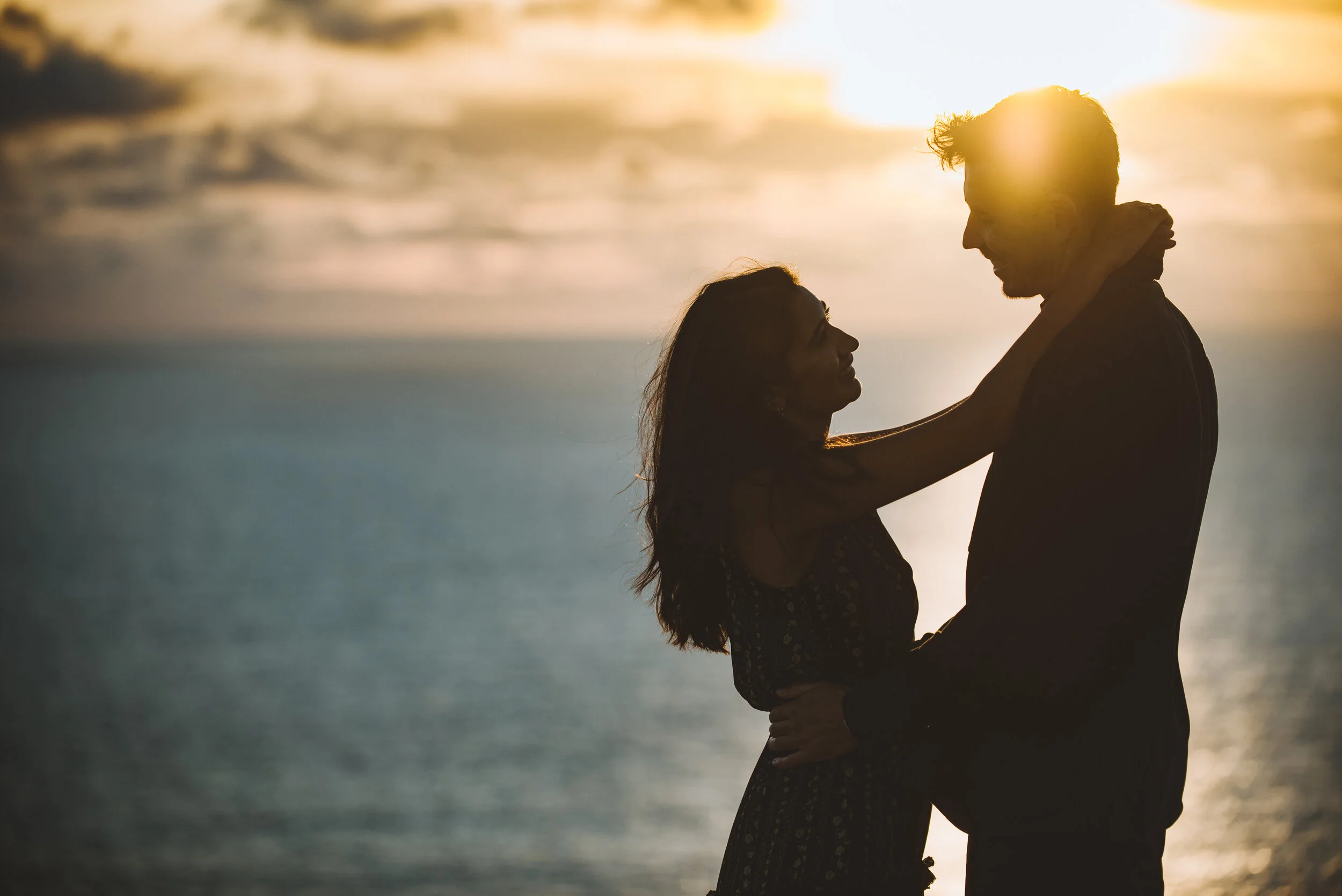 A couple embracing on a beach during sunset, with the woman looking up at the man.