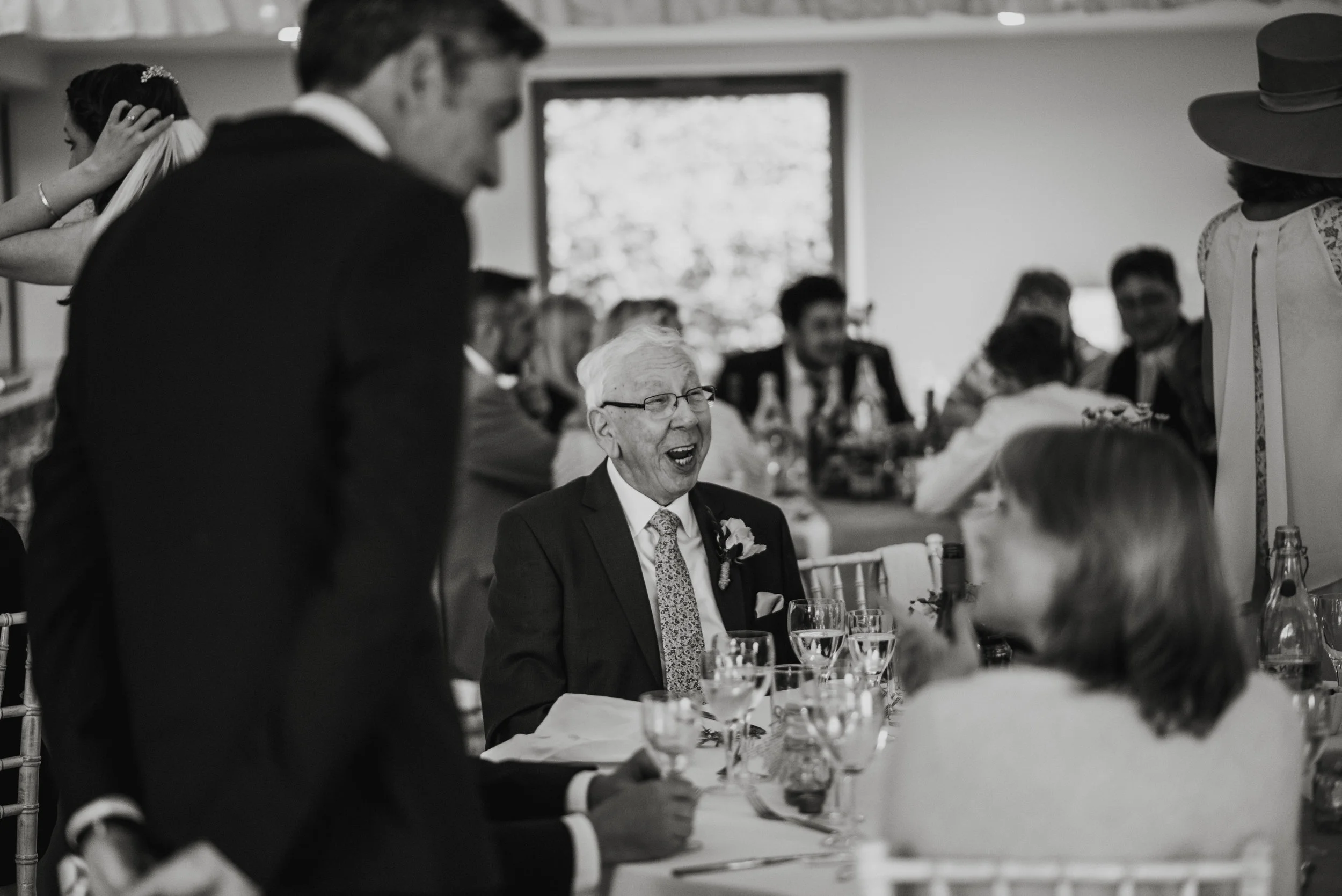 An elderly man in glasses and a suit with a boutonniere, smiling and talking at a formal gathering, with other guests seated at a long table, some standing and mingling, in a well-lit room.