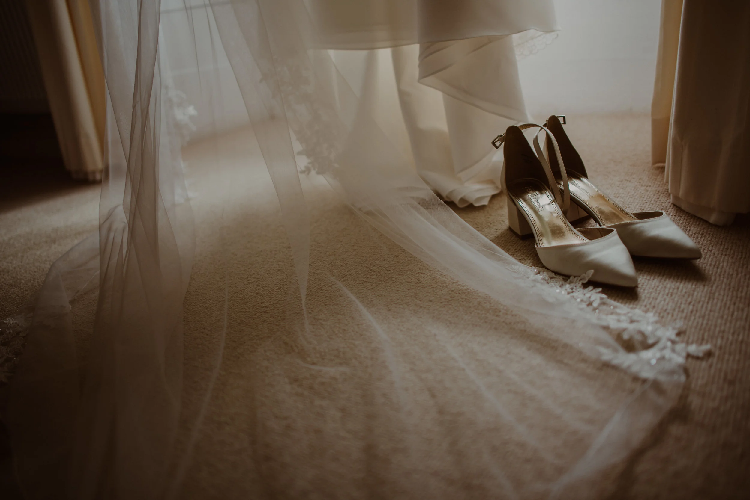 A pair of elegant white wedding heels placed on a beige carpet, with a wedding dress and sheer veil hanging above, illuminated by soft natural light.