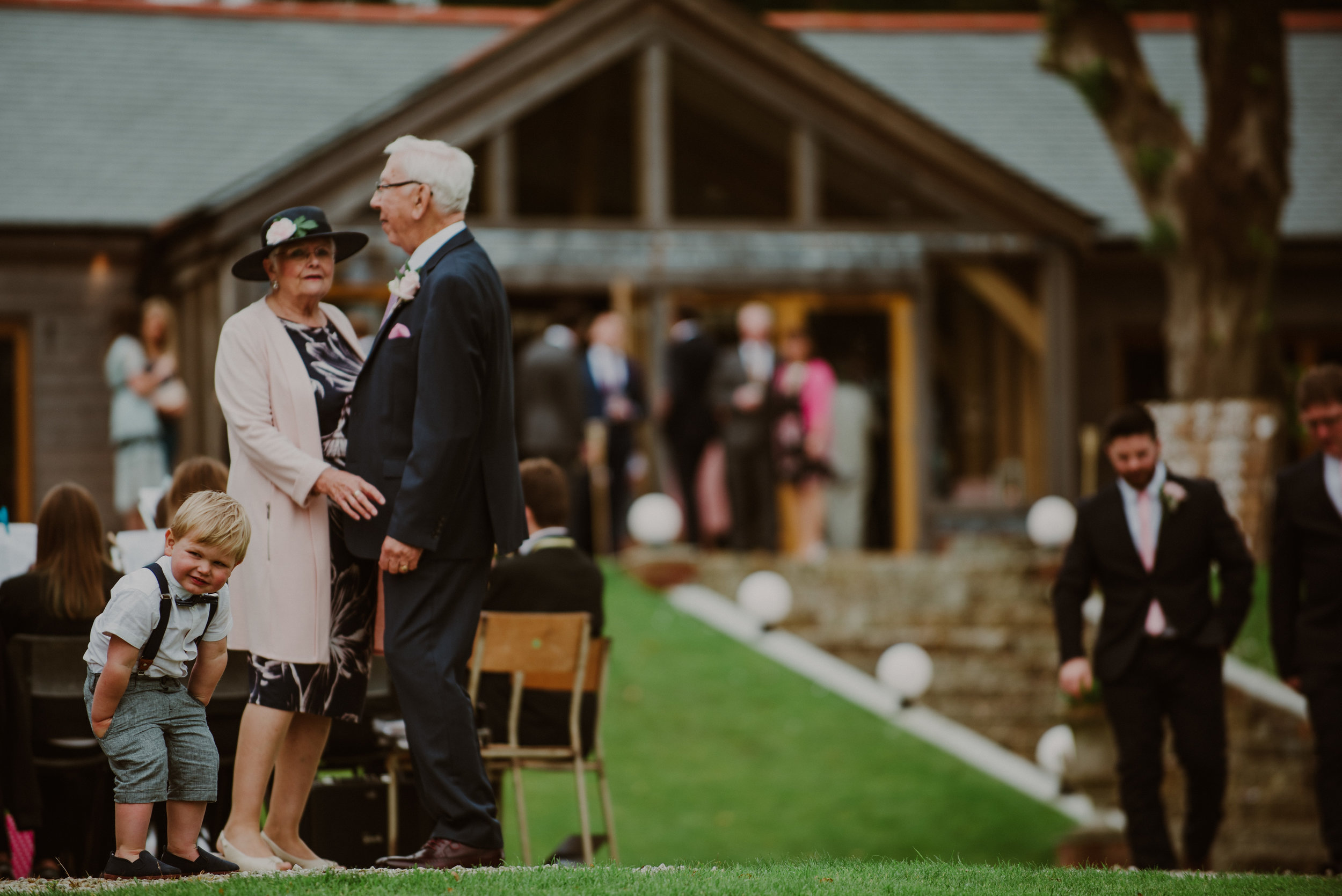 People at an outdoor wedding reception, including a young boy leaning on a chair and talking to an older woman and man, with other guests in the background near a rustic building.