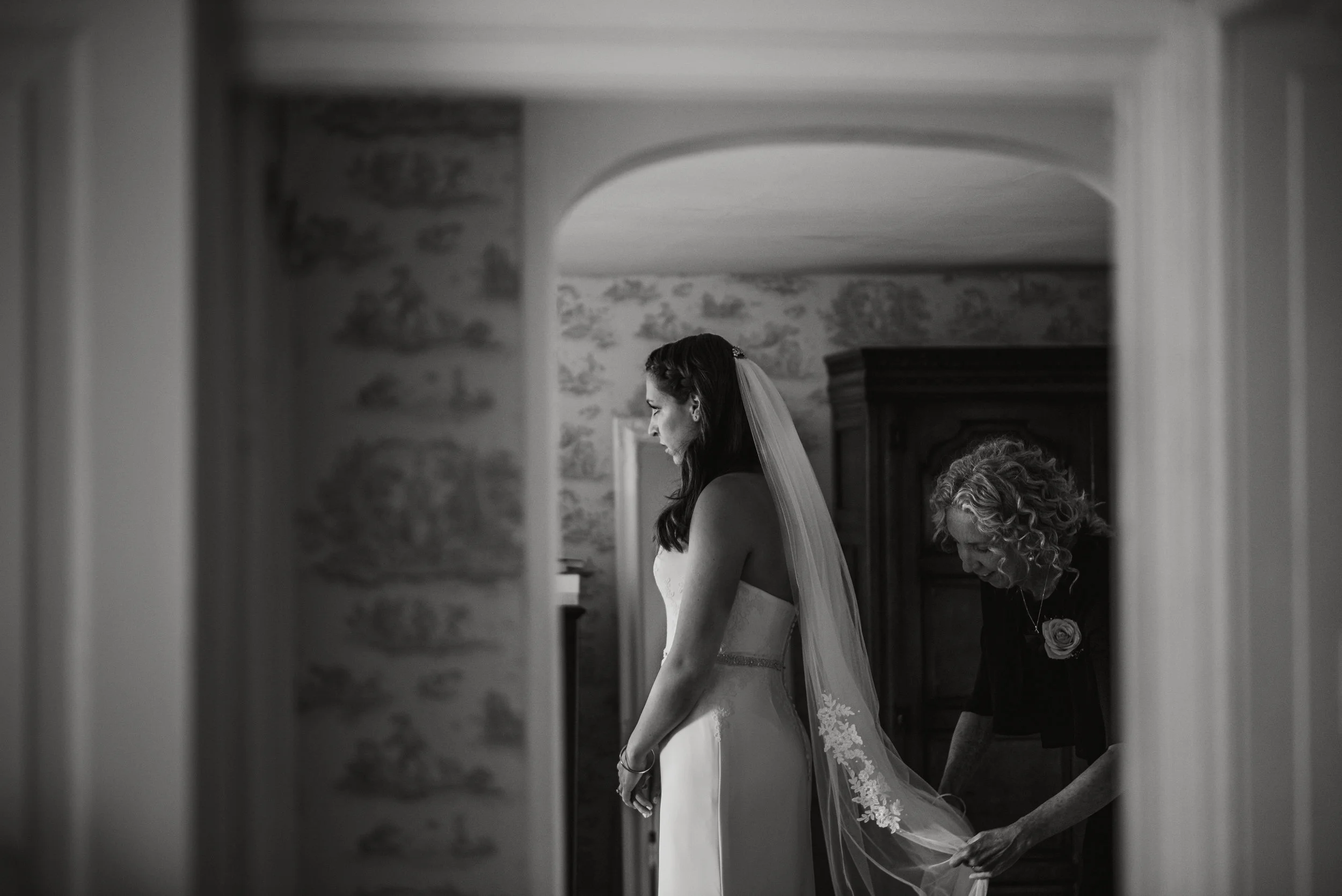 A bride wearing a strapless wedding dress with a jeweled waistband, her favorite veil, and a woman adjusting the veil from behind, in a room with vintage wallpaper and a wooden wardrobe.